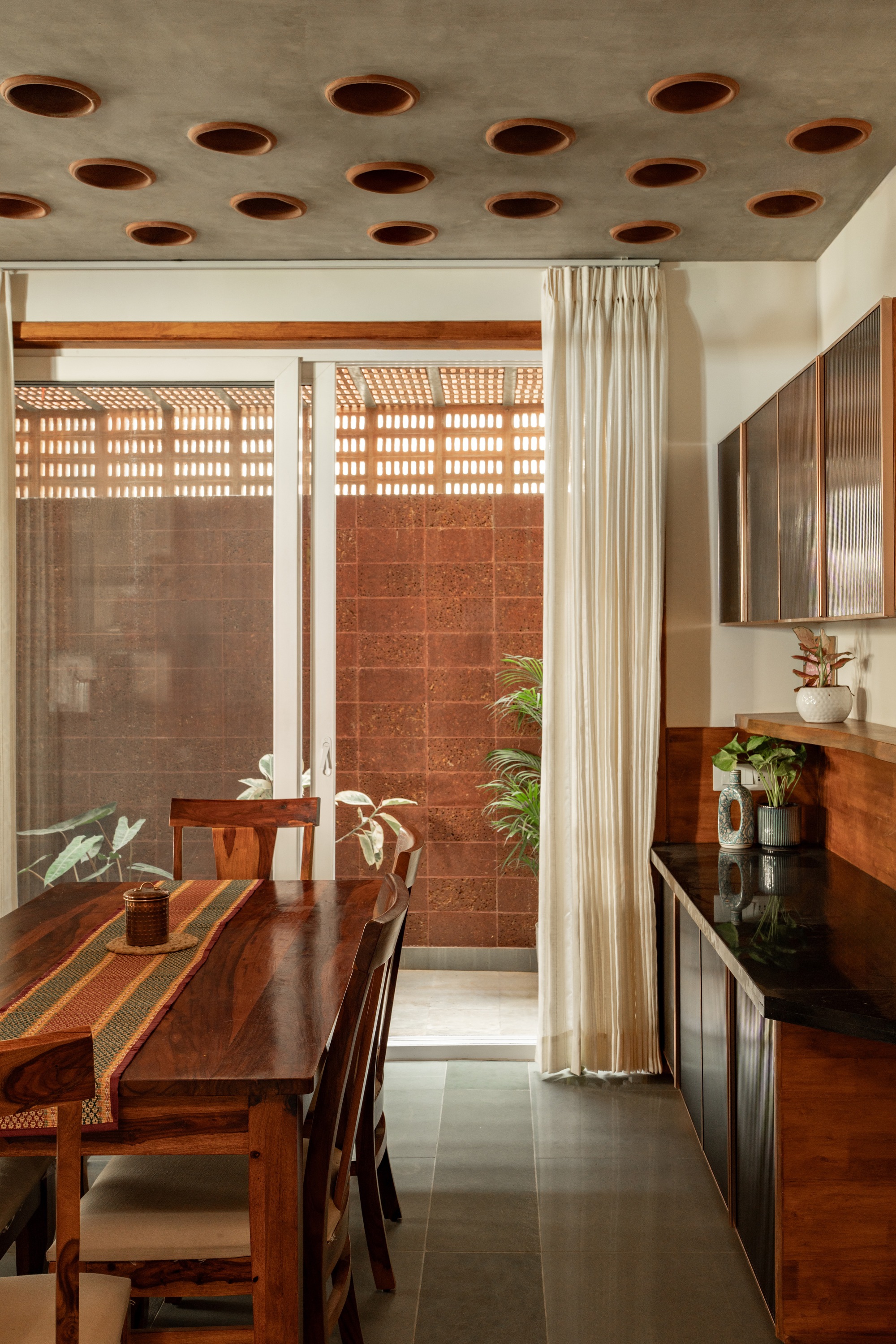 Dining area with perforated concrete ceiling opening to courtyard with terracotta tile wall and plants