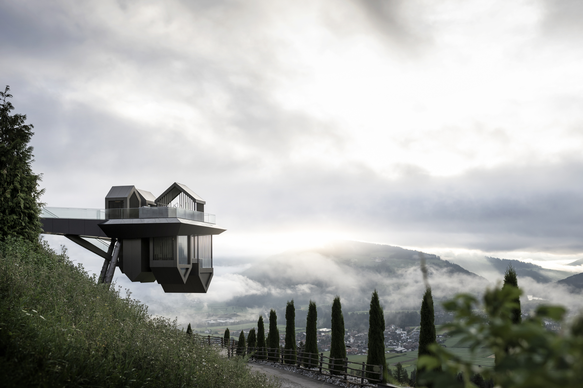 Cantilevered structure with gabled volumes suspended over a misty alpine valley lined with cypress trees