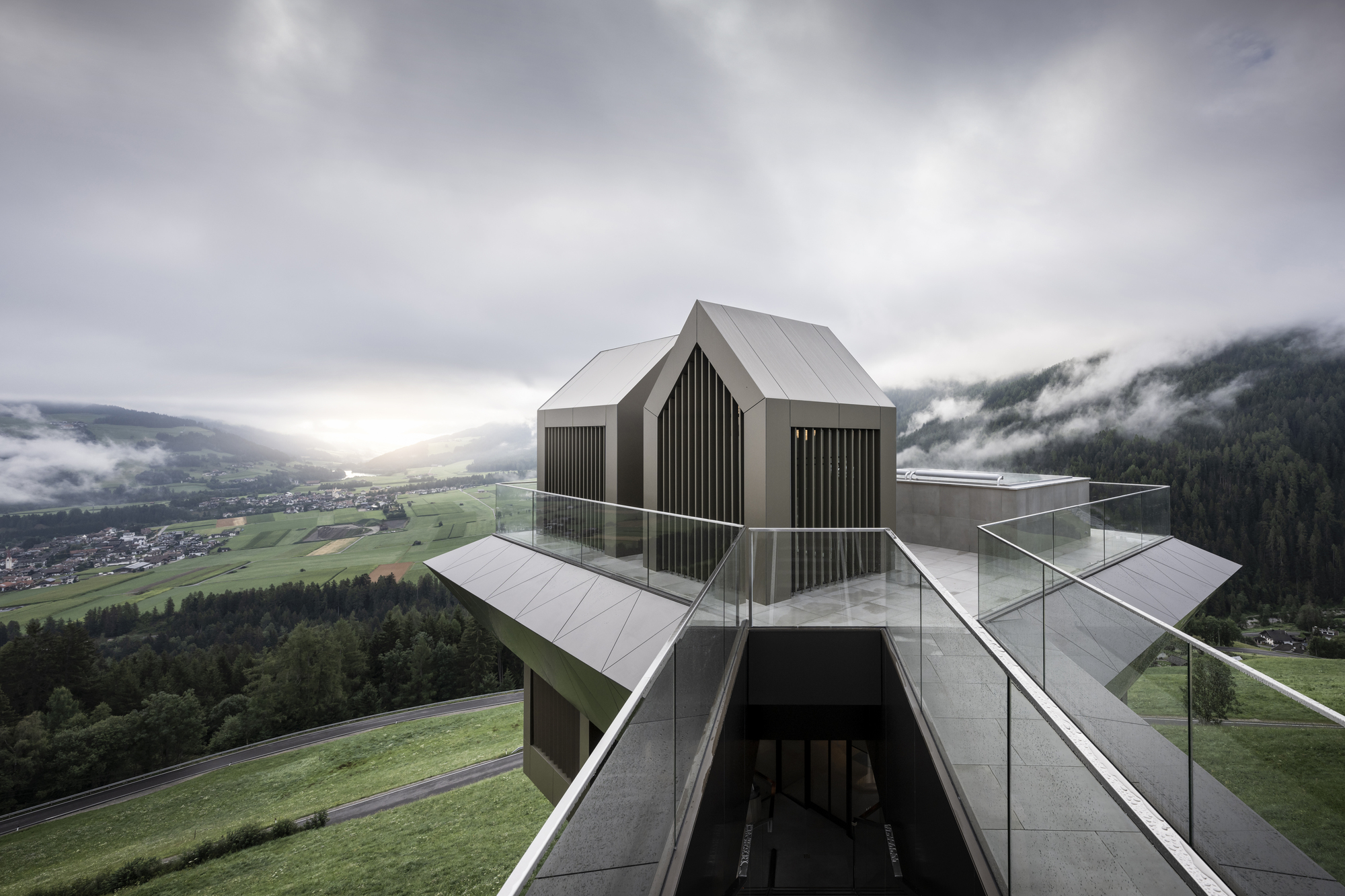 Three dark gabled volumes with vertical cladding atop a faceted metal base under heavy cloud cover