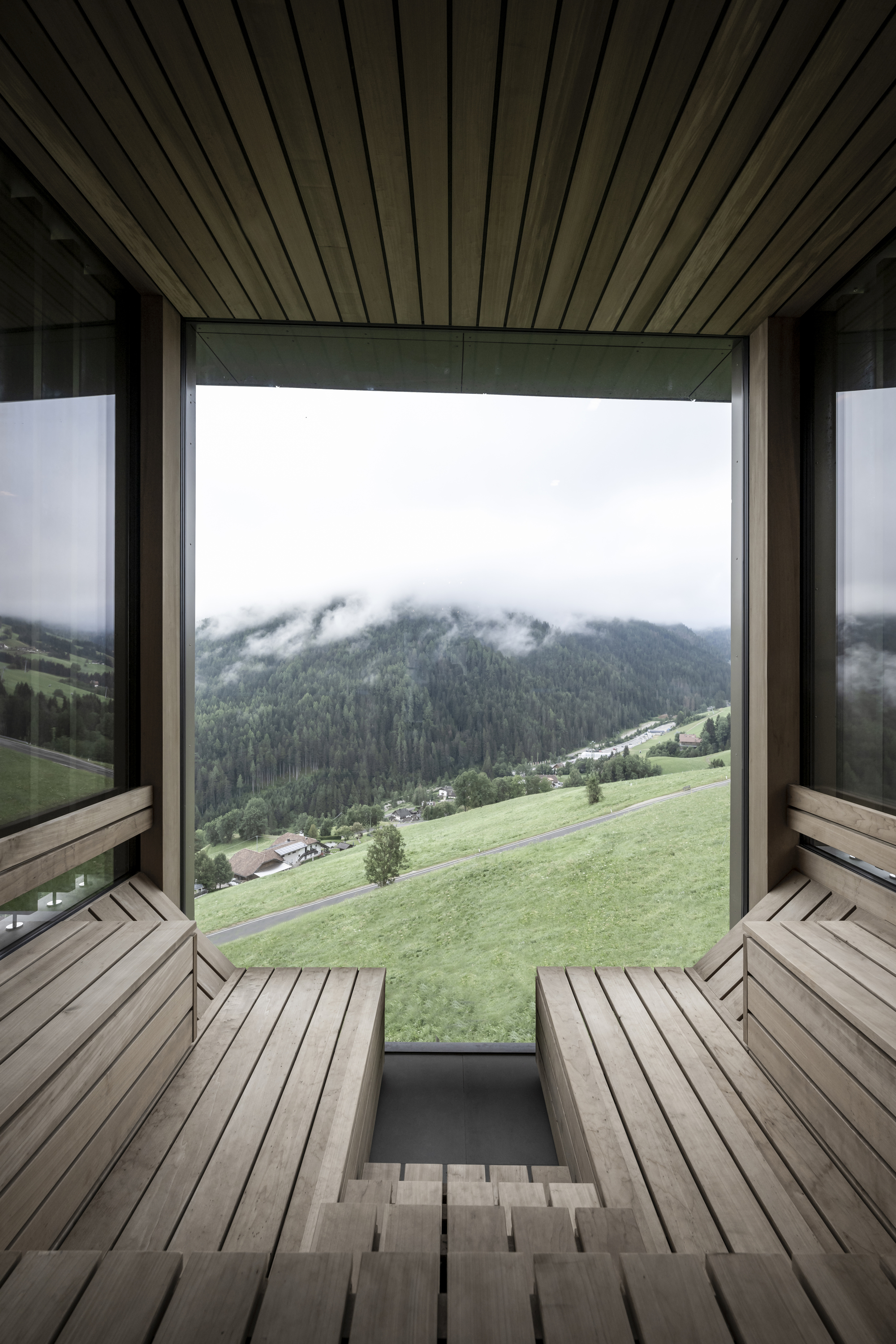 Timber sauna interior with slatted benches facing a picture window overlooking green meadows and mist