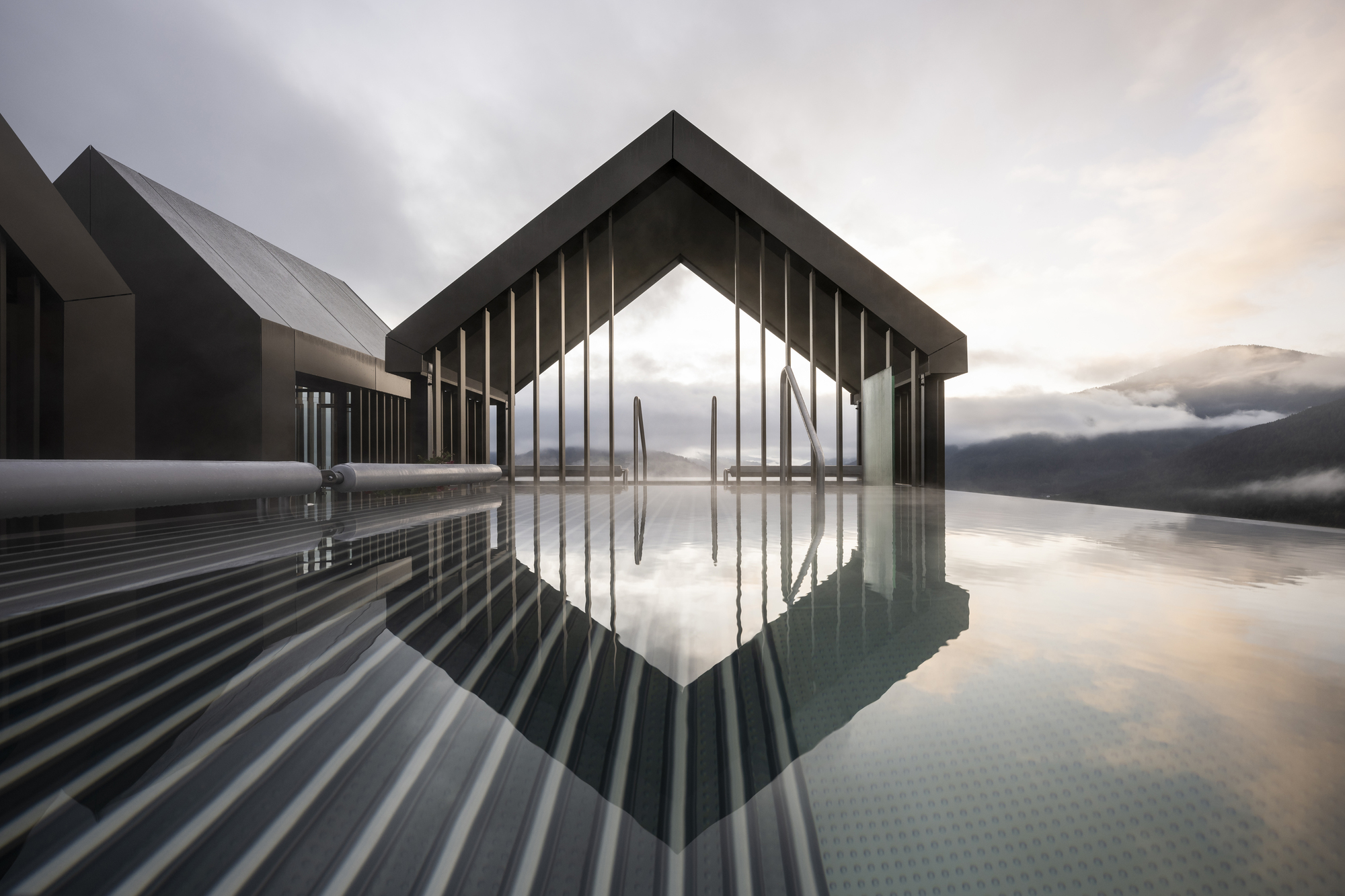 Gabled pavilion with vertical slat screen reflected in still water under overcast mountain sky