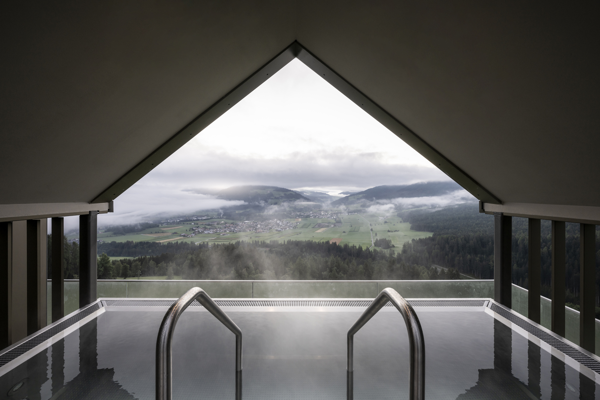 Indoor pool with house-shaped handrails framing view of misty valley and village below