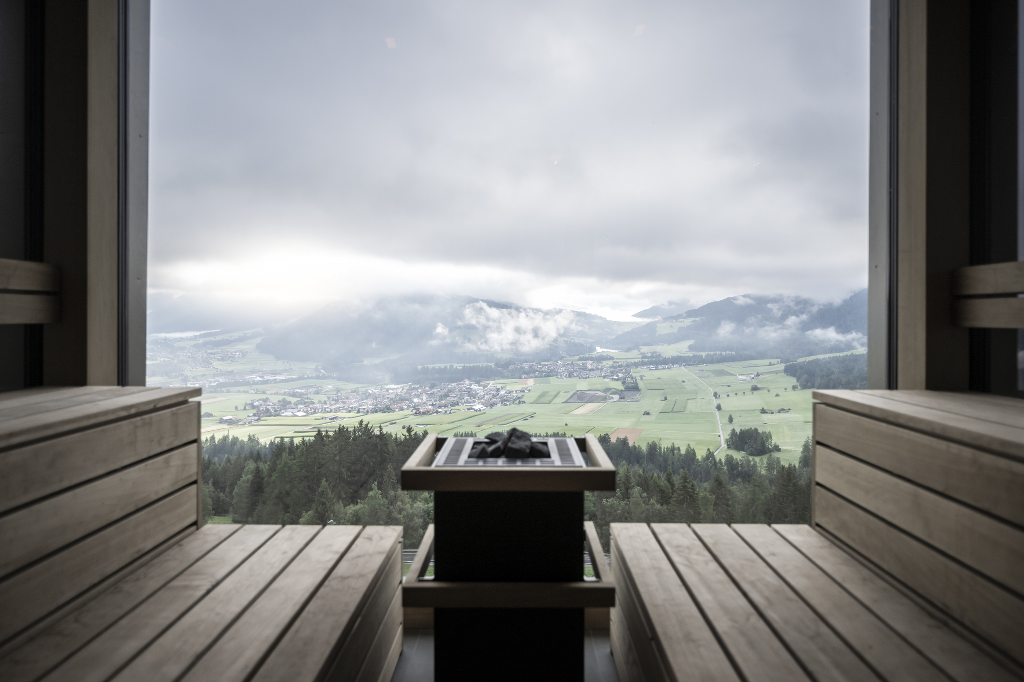 Sauna bench with heater and towel facing wide window overlooking patchwork fields and low clouds