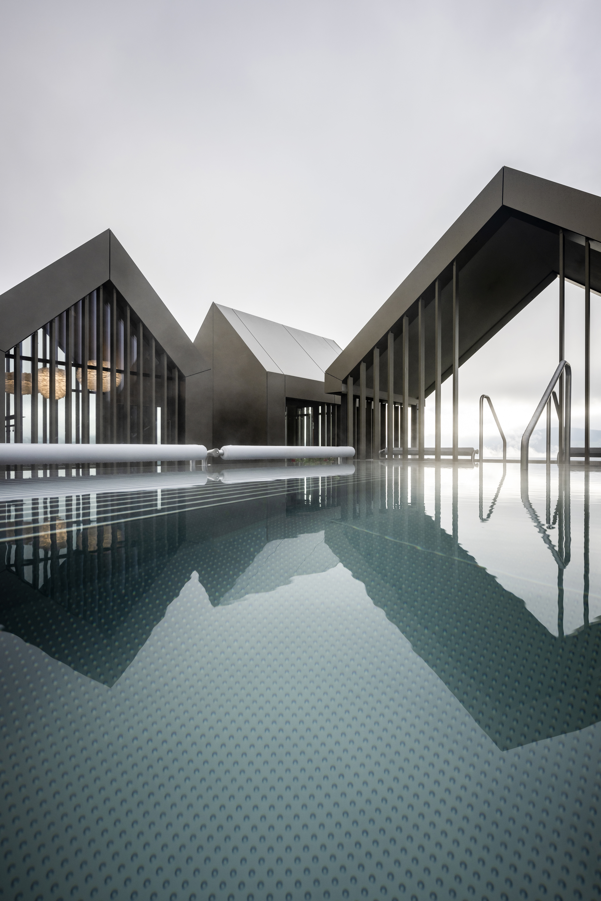 Rooftop pool with dark tile surface reflecting the angular gabled volumes under an overcast sky
