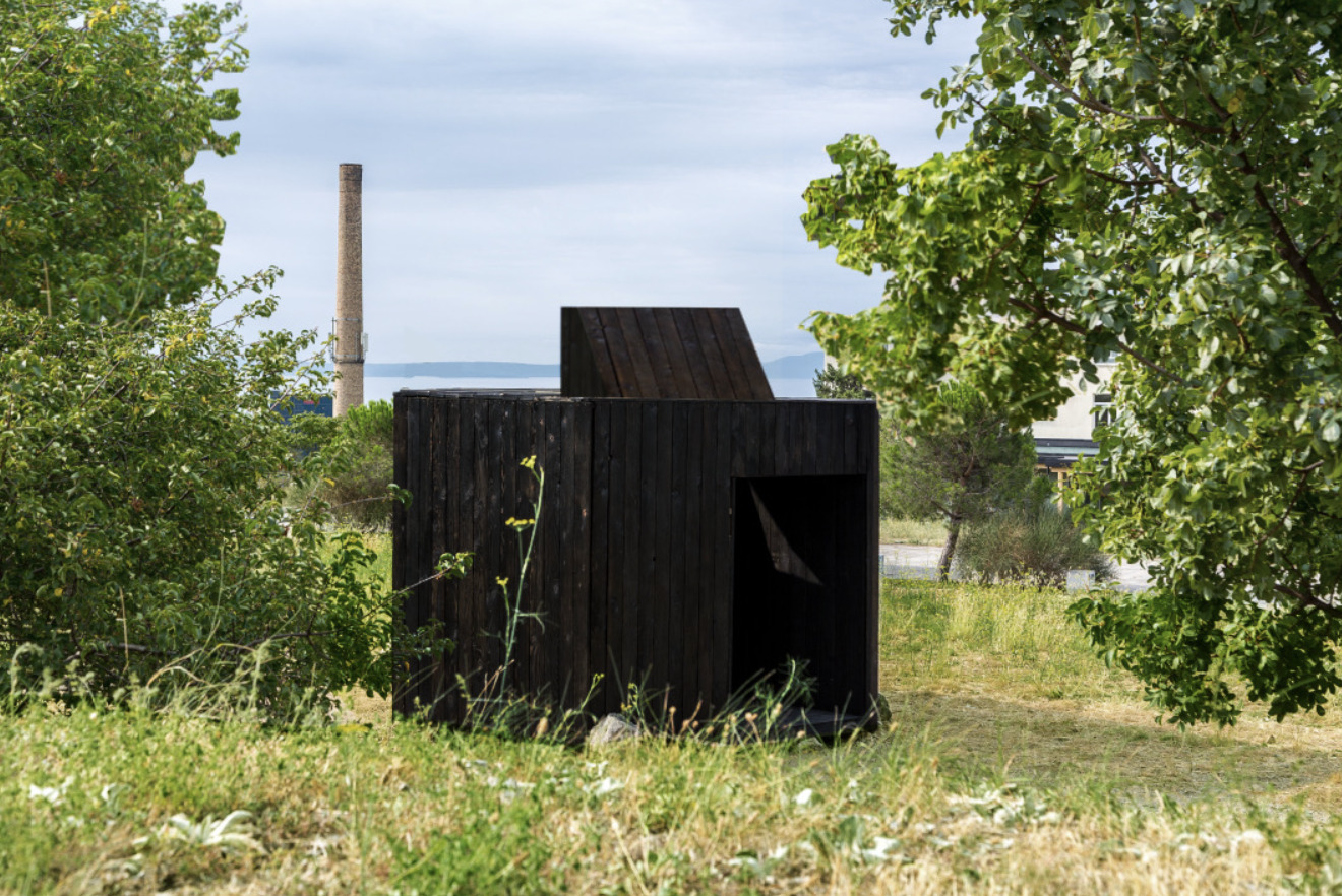 Charred timber structure with chimney and recessed openings framed by wild grasses and green foliage