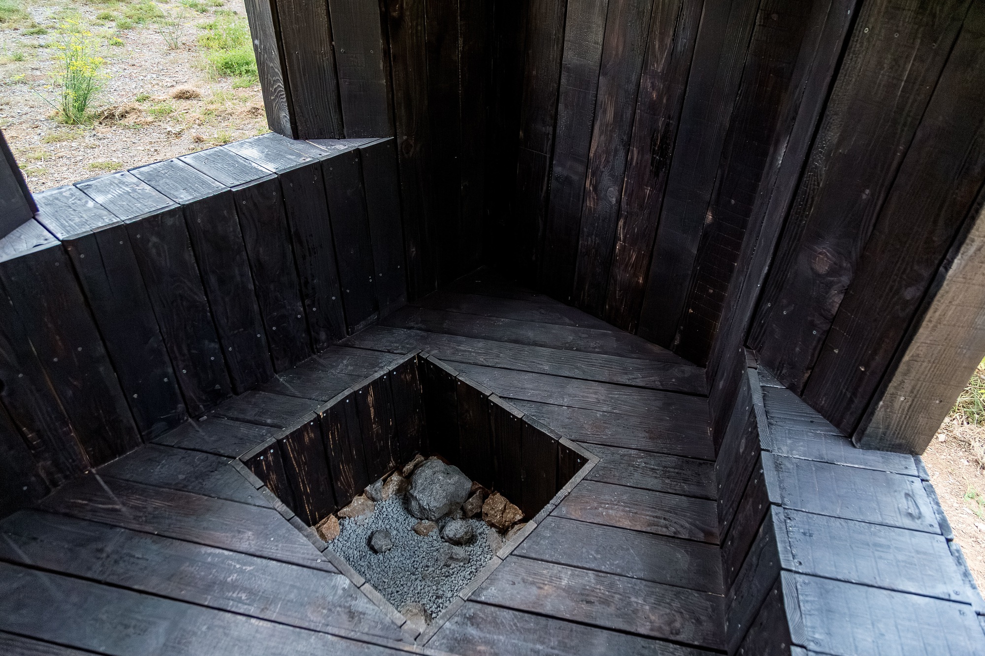 Top-down view of a square floor opening with stones below and charred timber bench surround