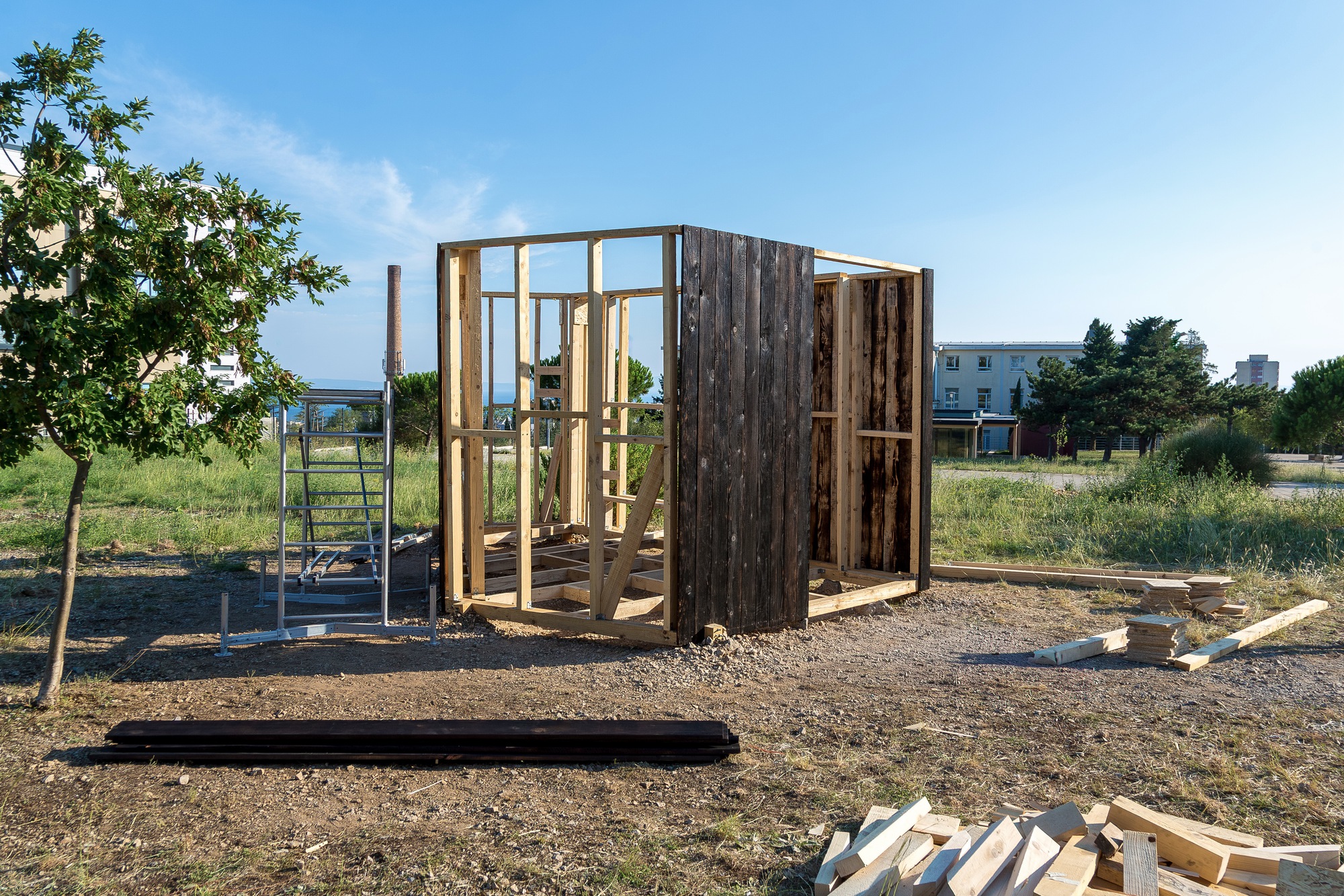 Timber frame structure under construction on a gravel site with exposed studs and weathered cladding panels