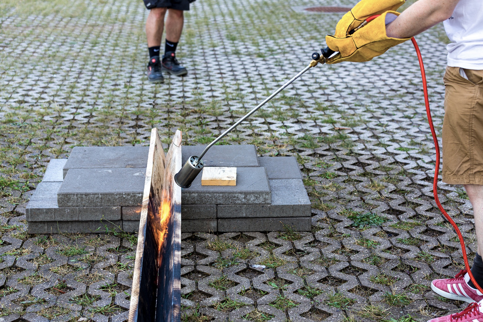 Hands using a torch to char timber samples arranged on concrete pavers in a plaza