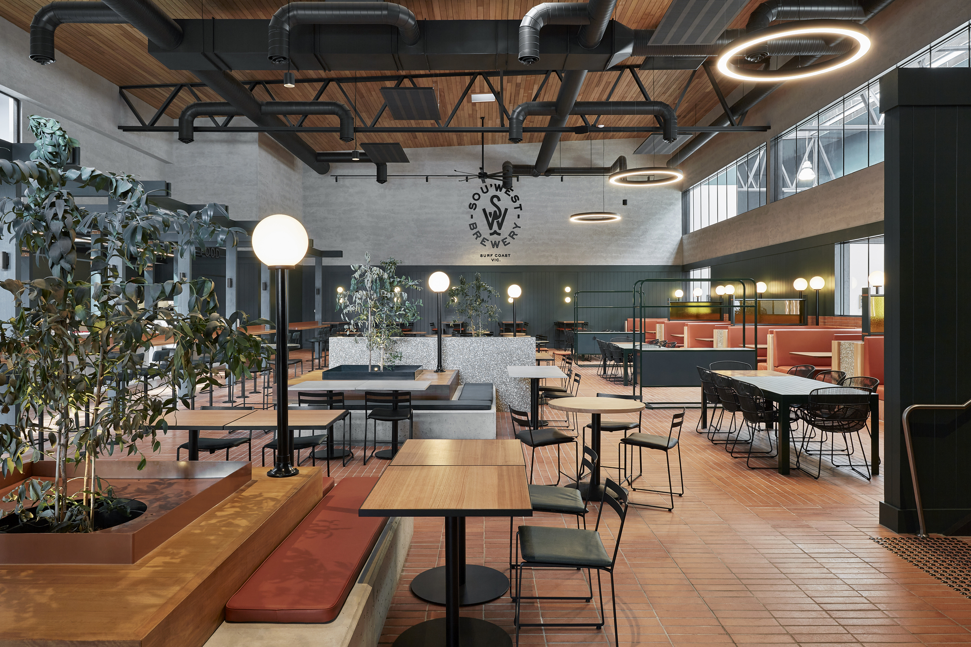 Taproom interior with exposed black steel trusses, timber ceiling panels and potted trees beside terracotta tile floor