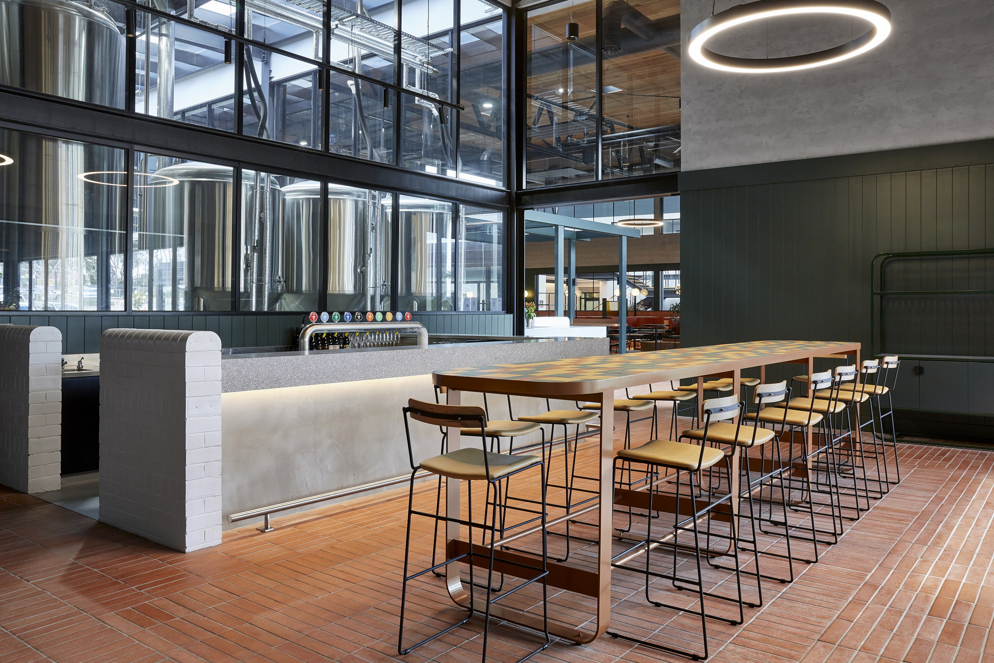 Brewery bar with high-top table and stools facing stainless steel fermentation tanks behind glazed partition