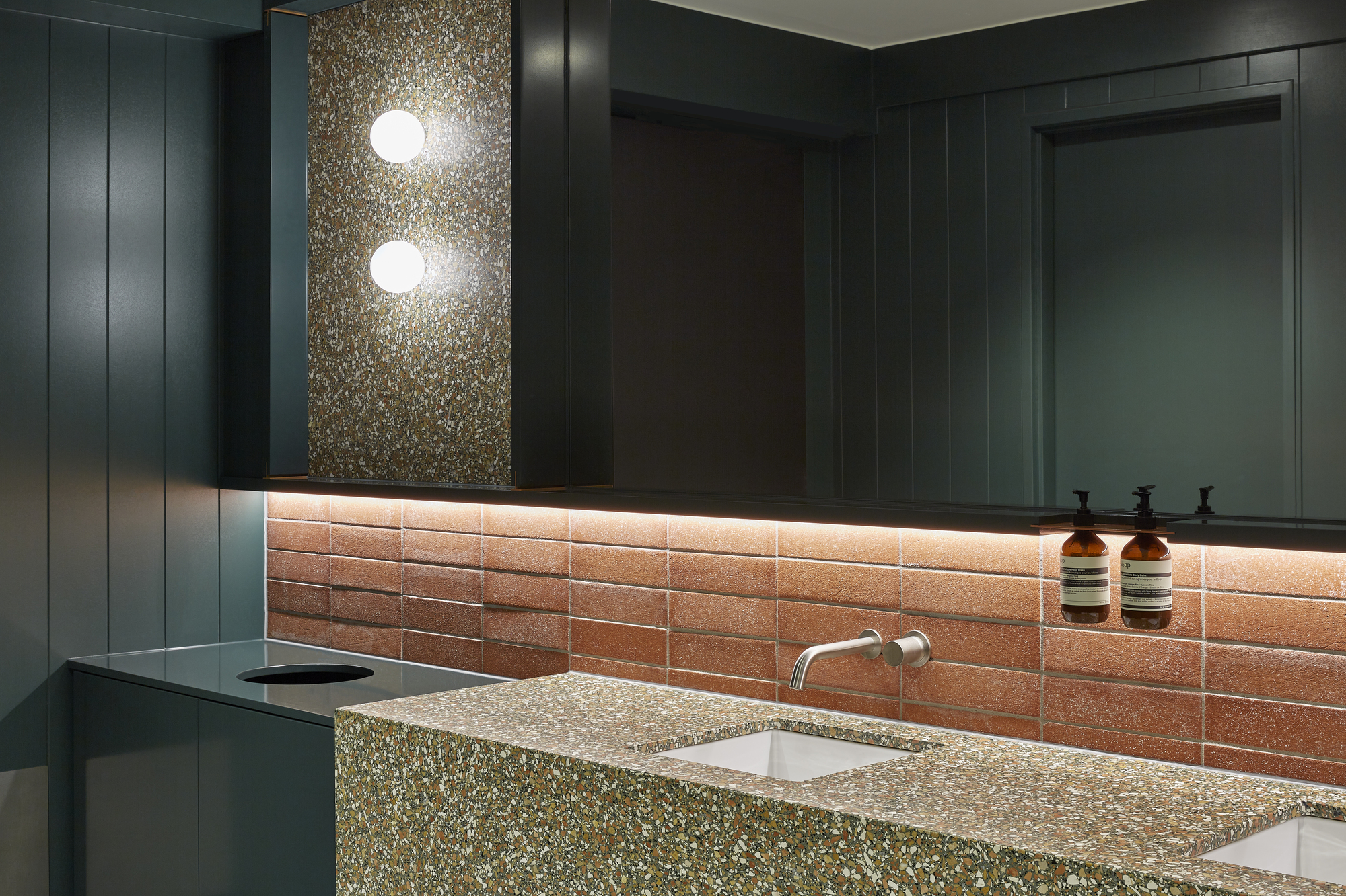 Washroom with terrazzo countertop, terracotta subway tiles, and dark paneled walls with linear lighting