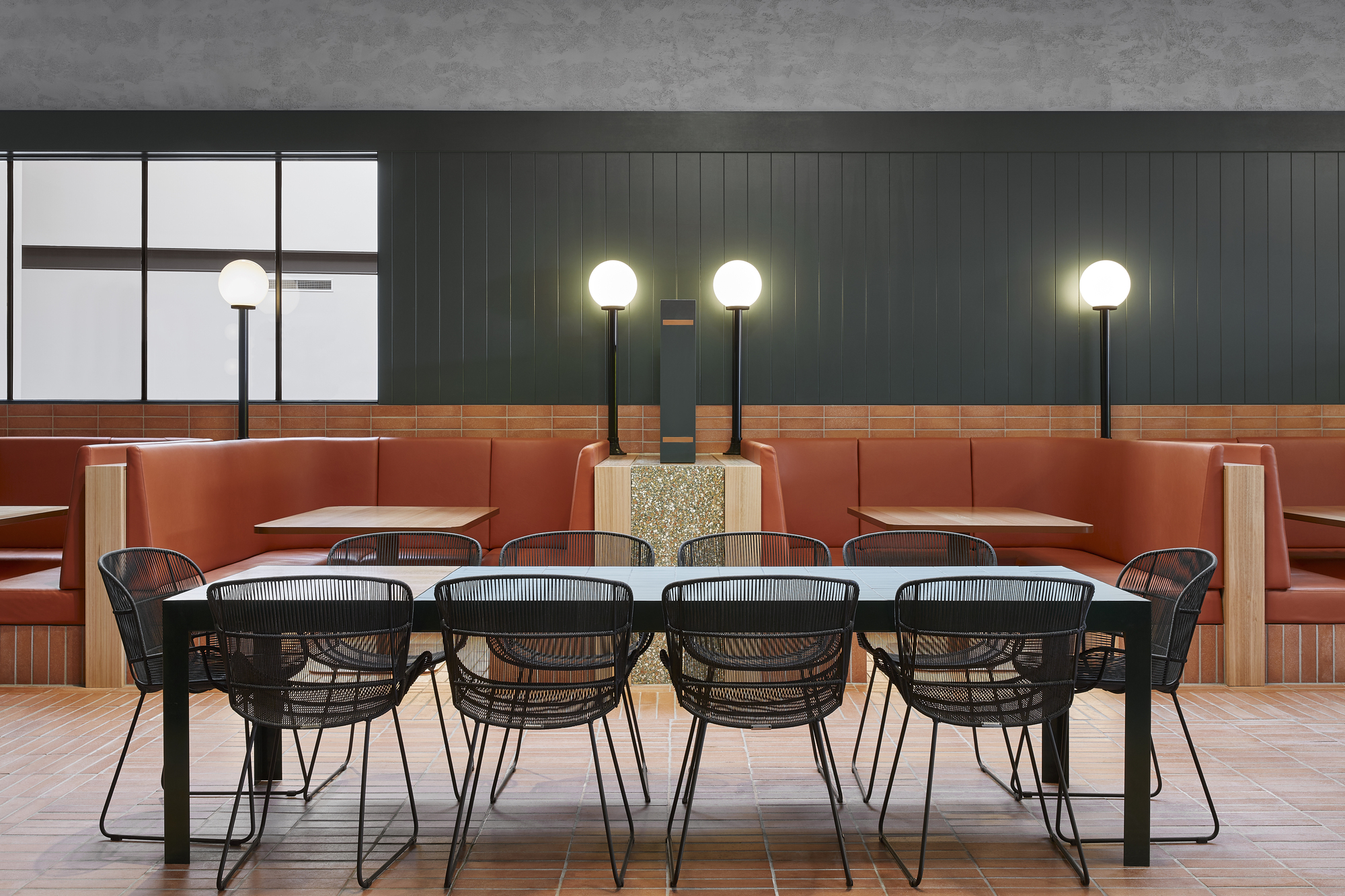 Communal dining table with wire chairs and terrazzo plinth beneath globe pendant lights at dusk