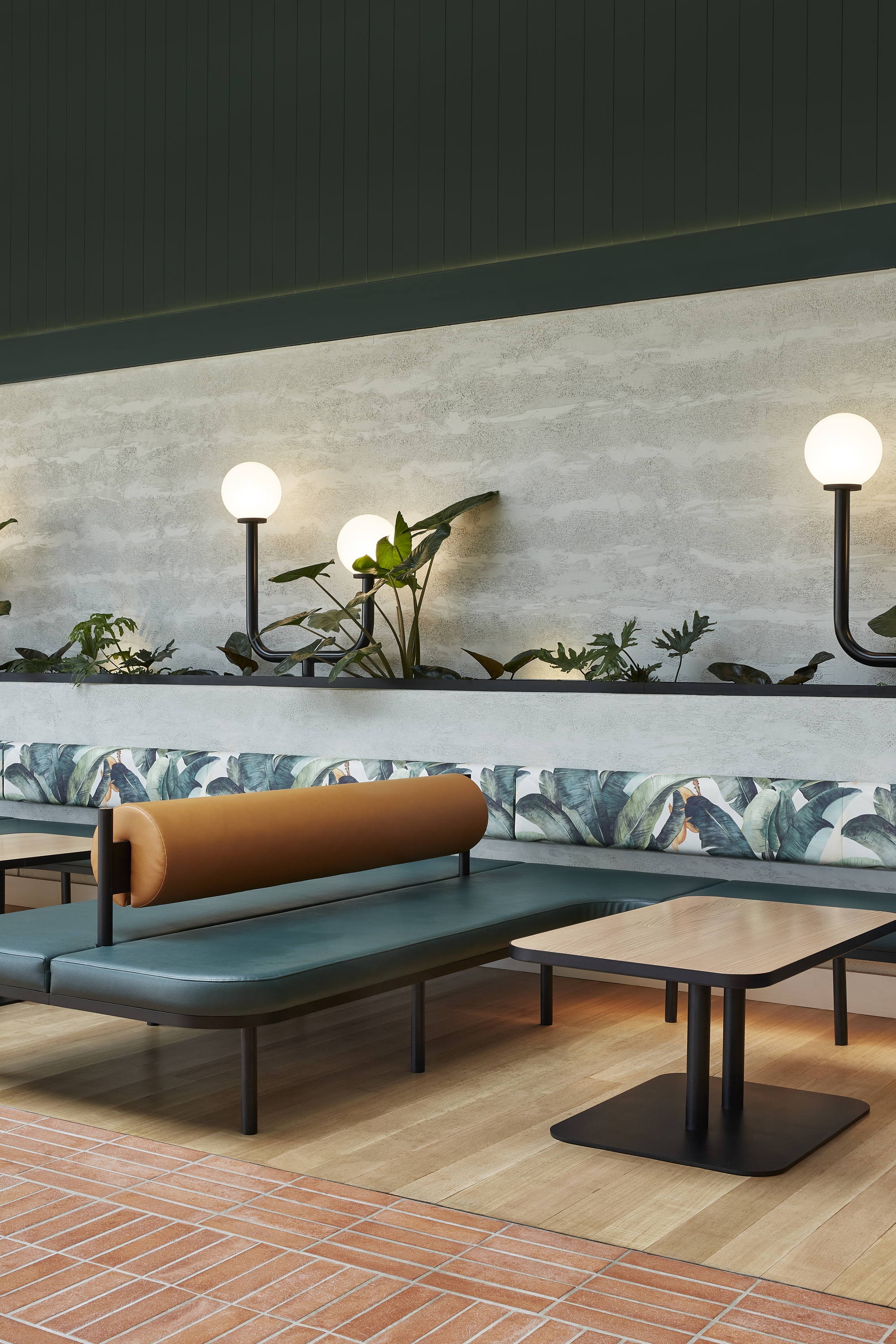 Banquette seating with leaf-print upholstery beneath a planted shelf with globe sconces and concrete wall