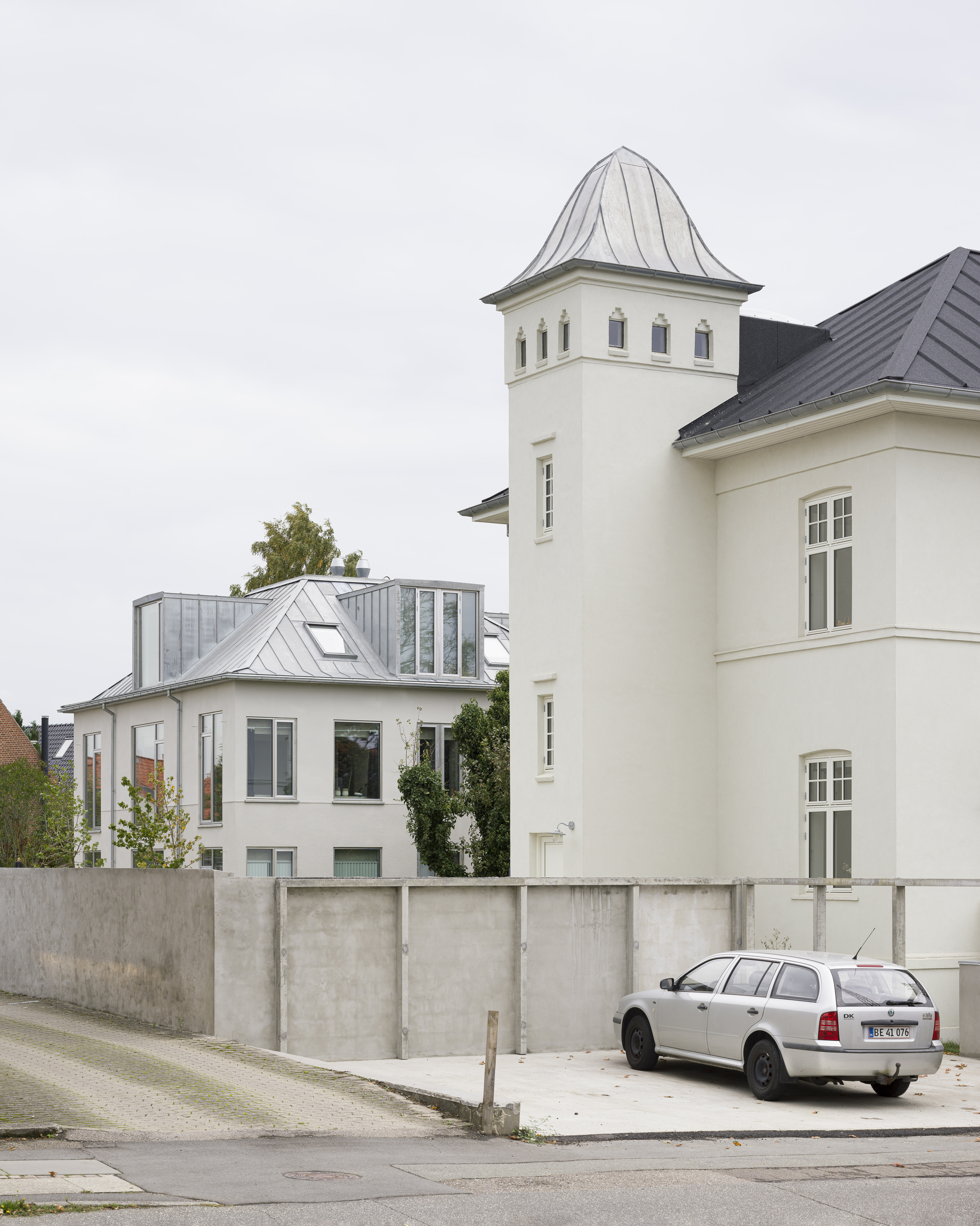 Street view of white tower with pointed roof beside zinc-clad addition behind concrete retaining wall