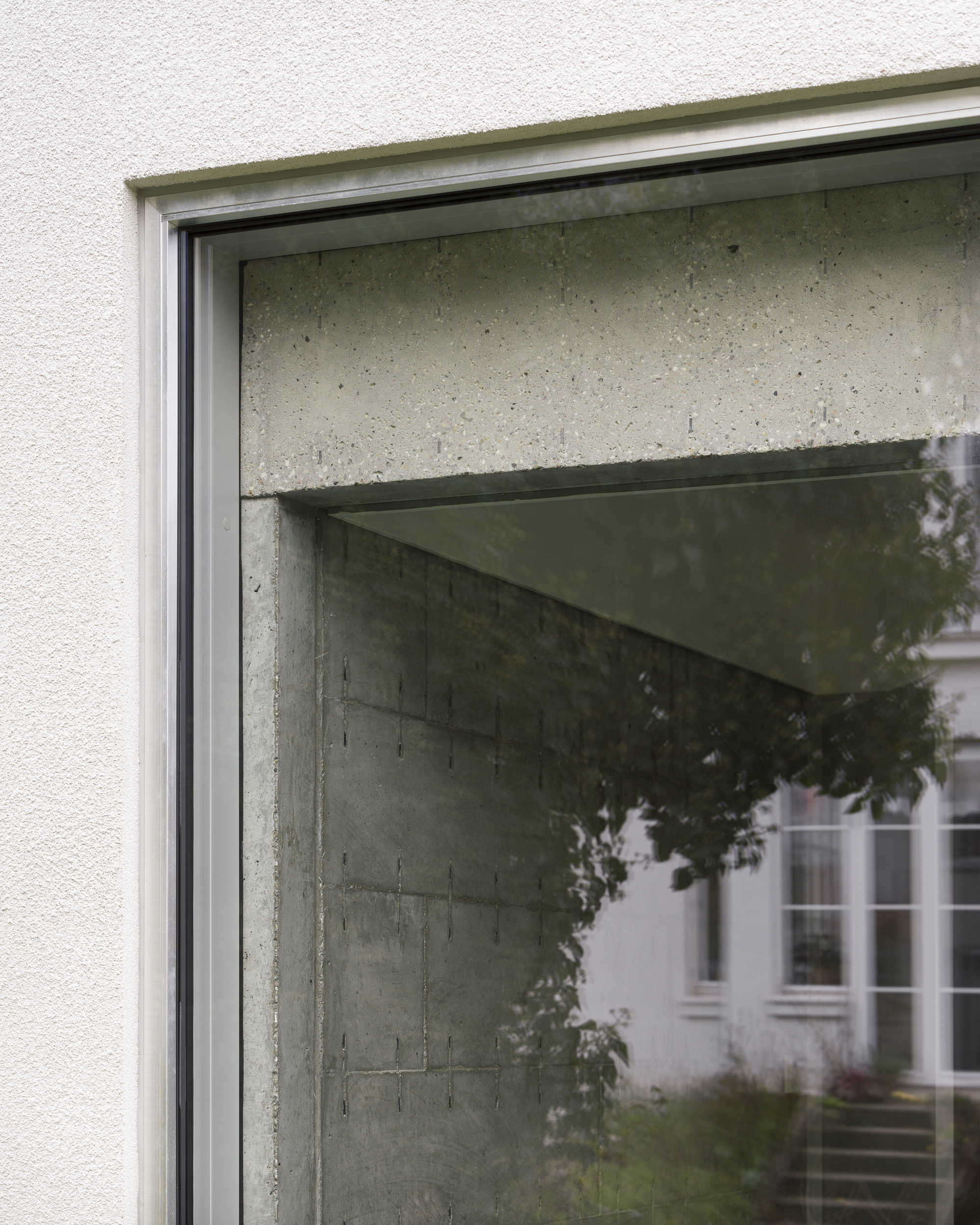 Window detail showing concrete lintel meeting plaster facade with reflected foliage in glass