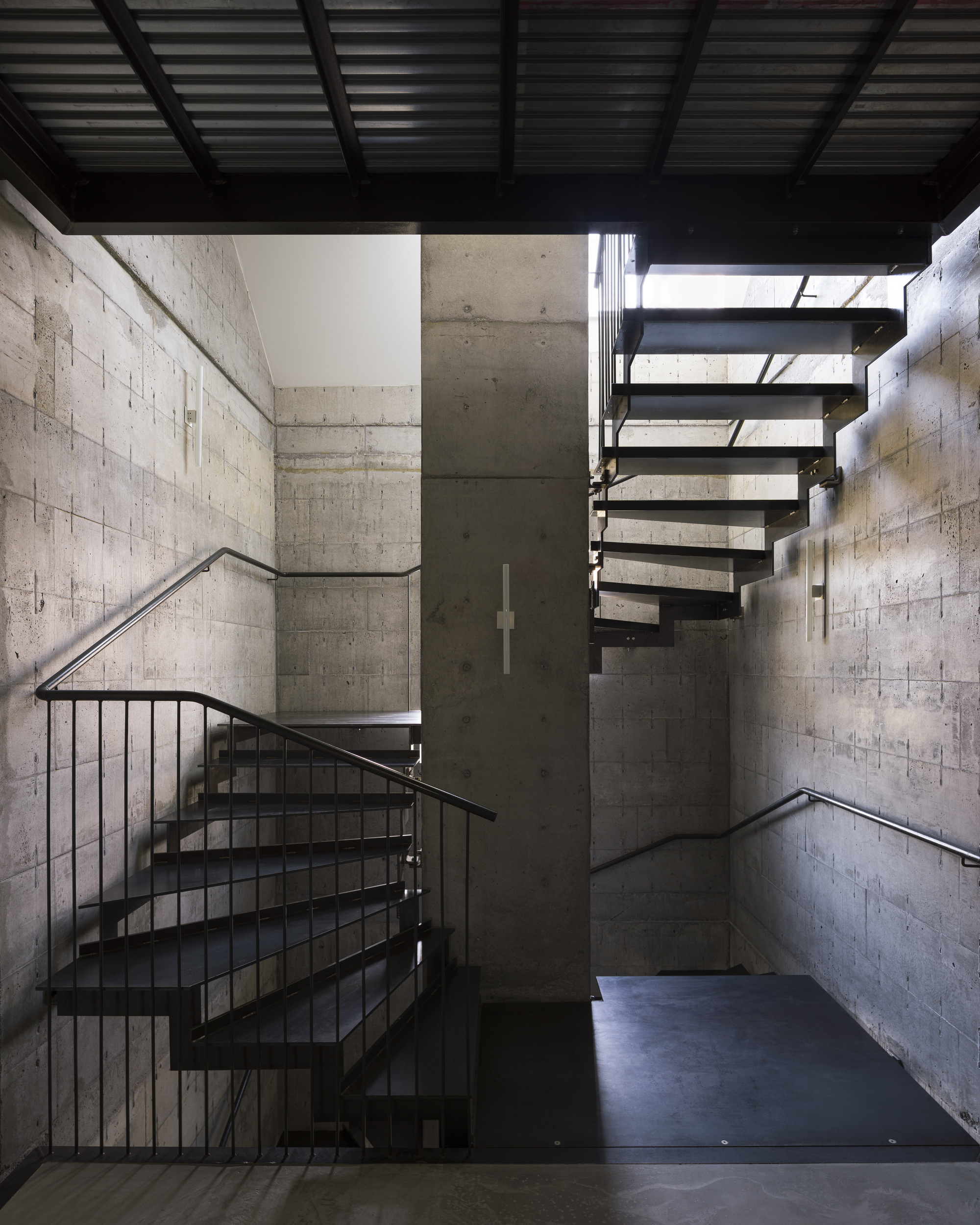 Double-height stairwell with exposed concrete block walls and black steel open-riser stairs under skylights