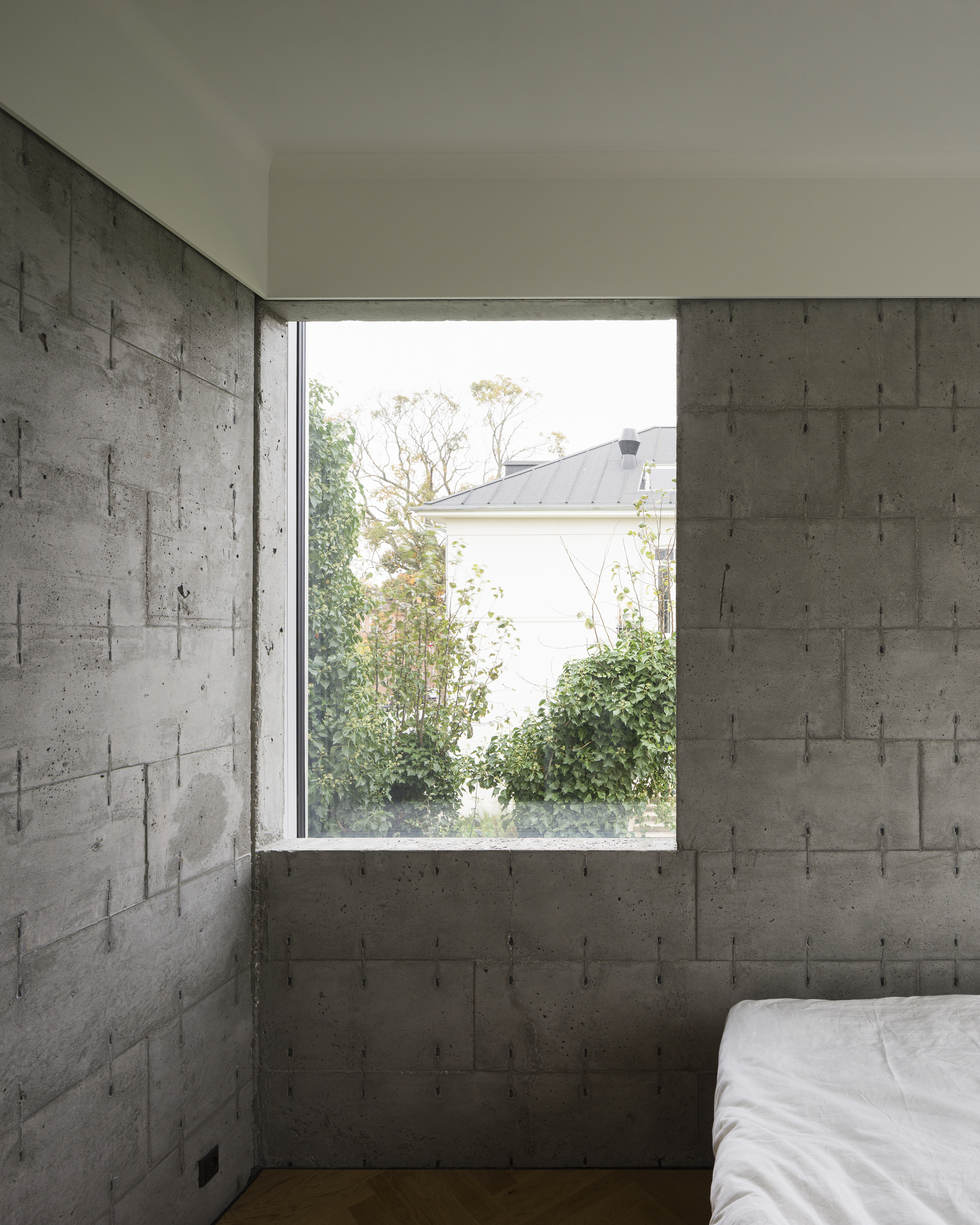 Bedroom corner with board-formed concrete walls and window overlooking trees and neighboring rooftop
