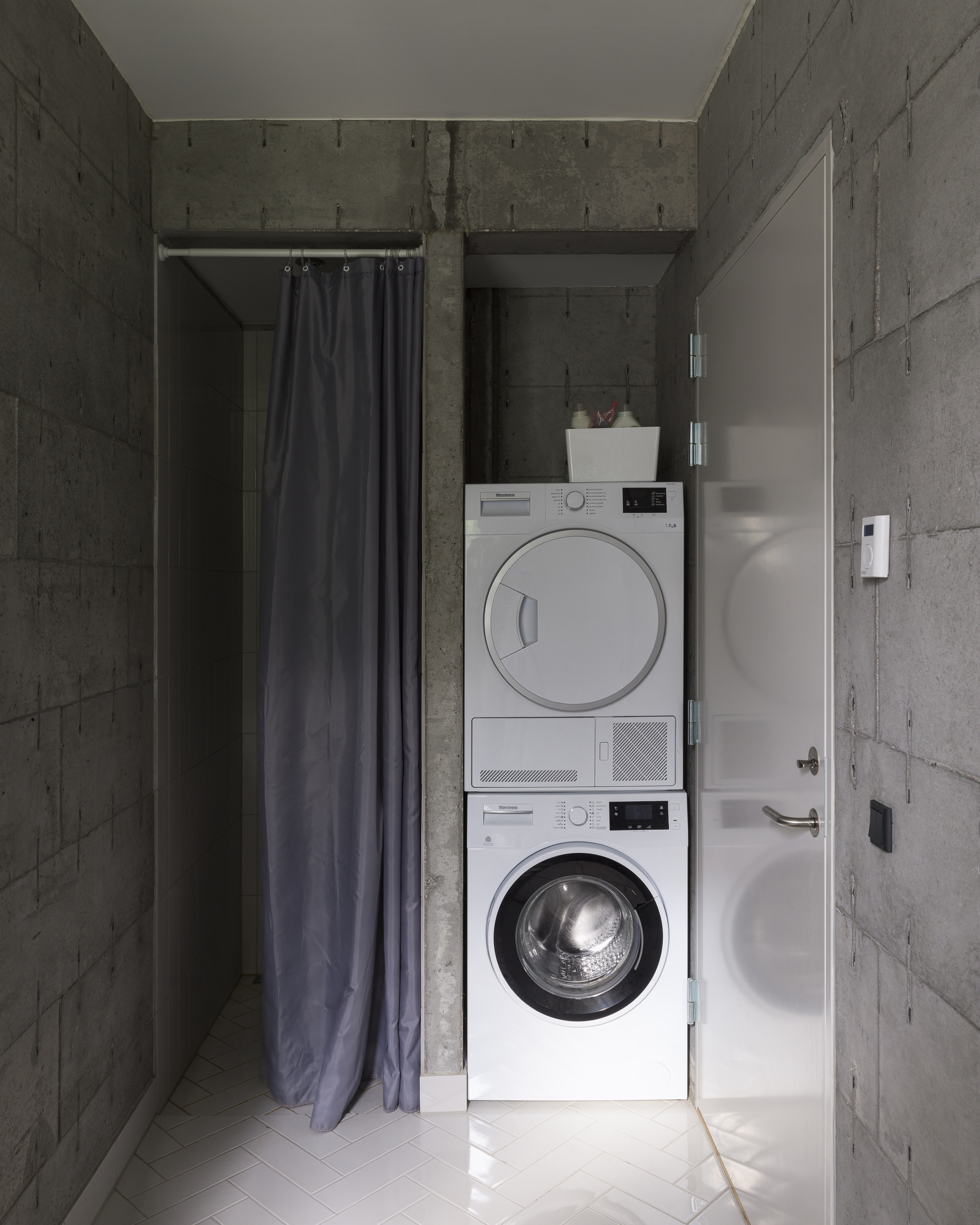 Compact laundry alcove with stacked washer and dryer flanked by dark curtains and concrete walls