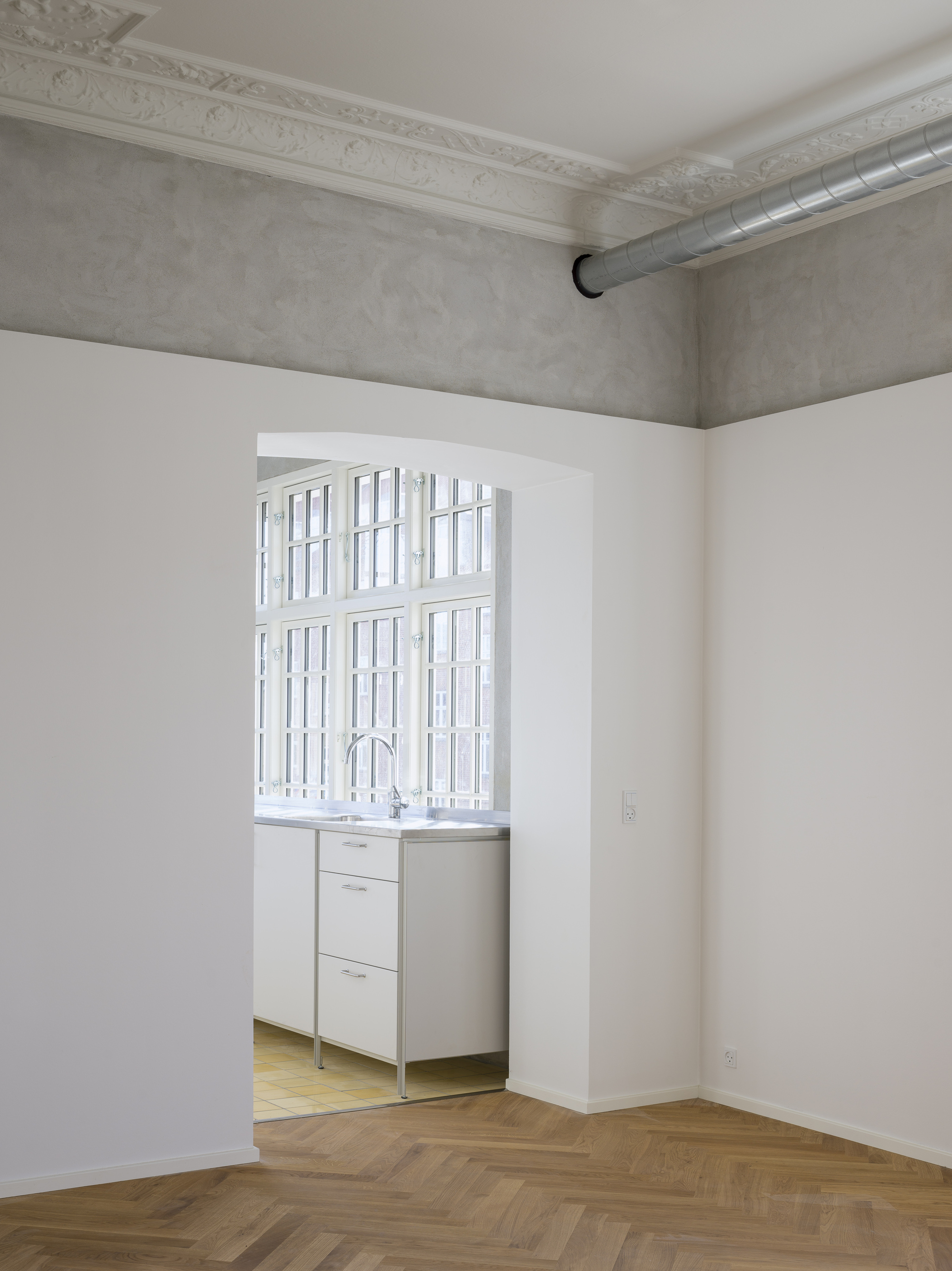 View through white-walled rooms with herringbone wood floors toward a kitchenette beneath gridded casement windows