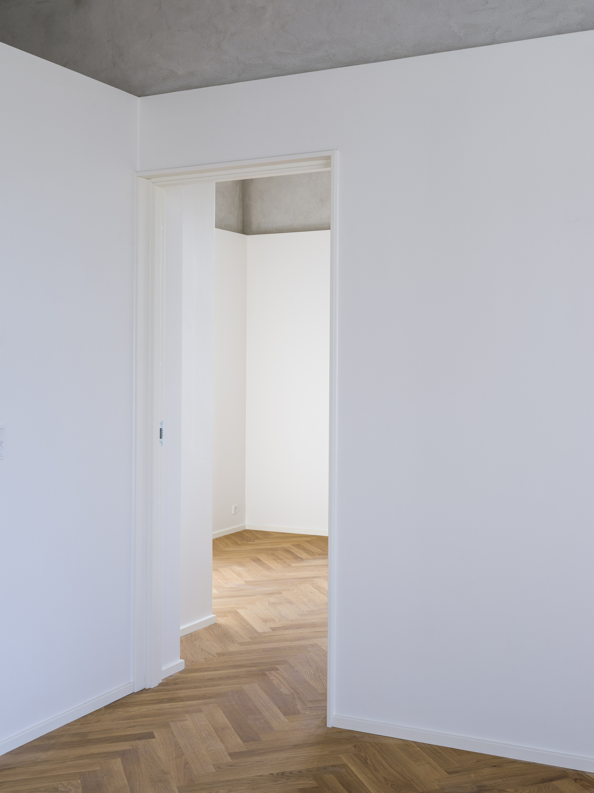 Doorway framed in white trim connecting rooms with herringbone parquet and exposed concrete ceiling beam