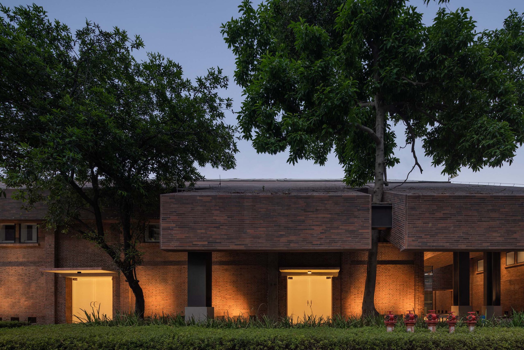 Brick colonnade with interior lighting at dusk, framed by mature trees