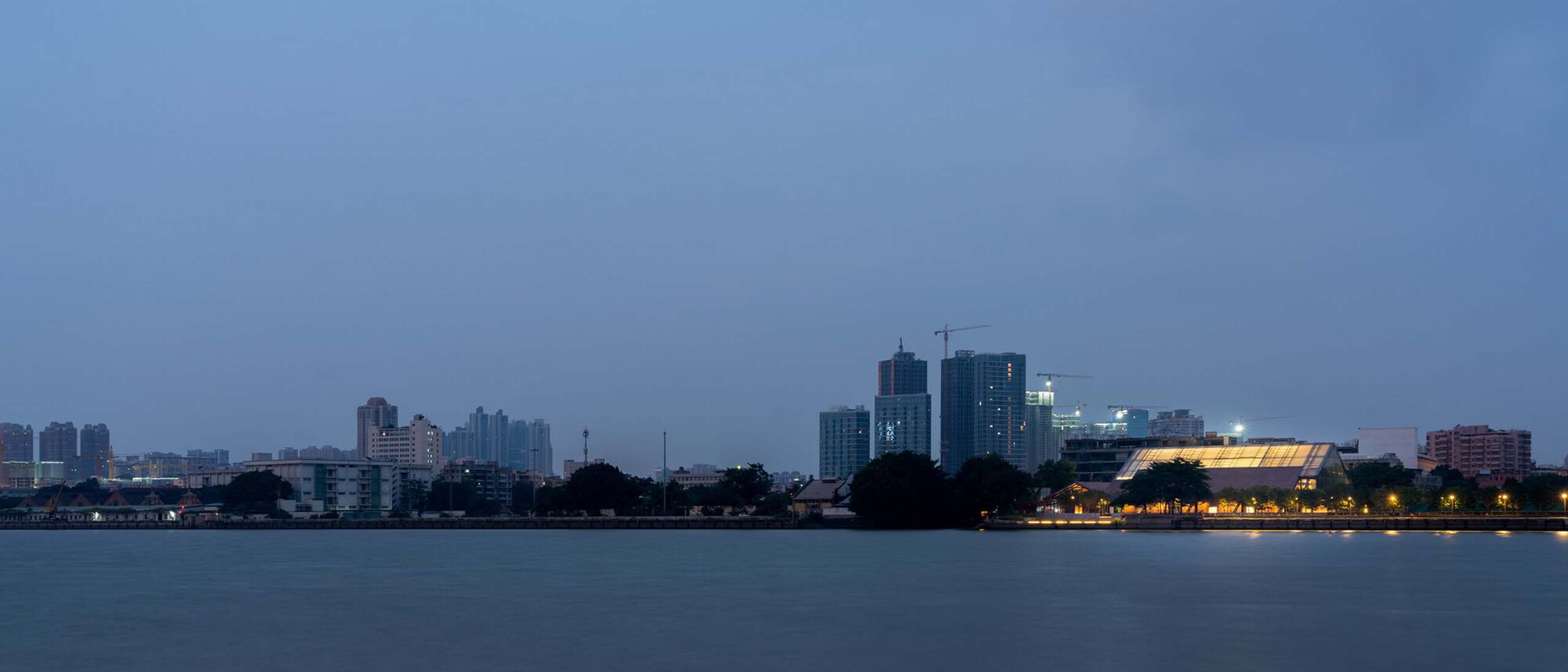 Distant view of the illuminated riverside structure at twilight with urban skyline behind