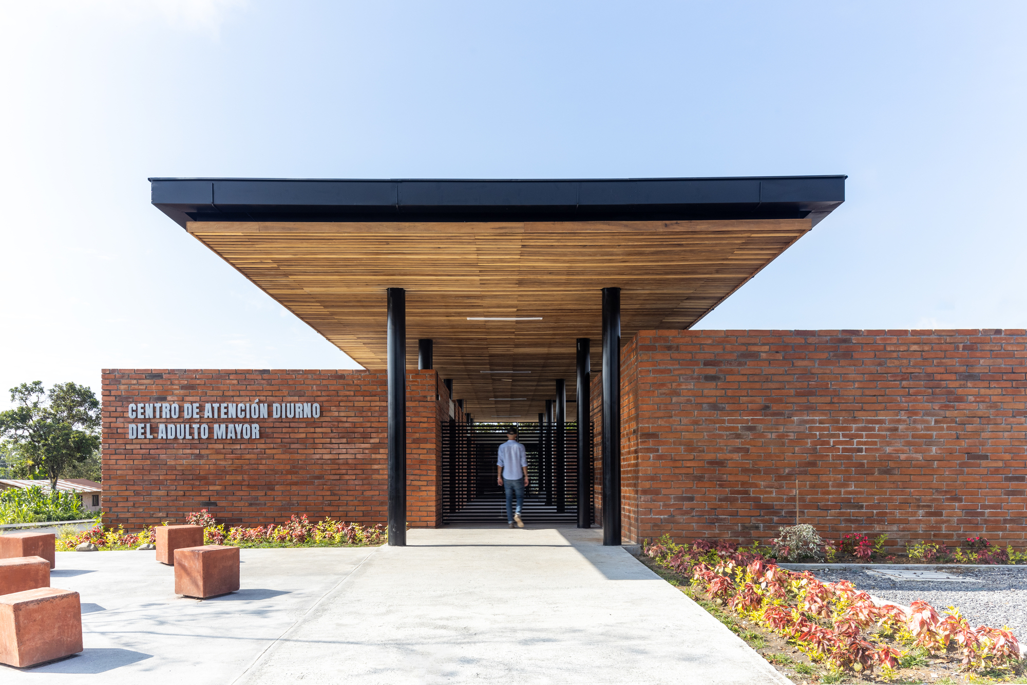 Entry corridor with timber slat ceiling and black steel columns framing a figure walking toward the courtyard