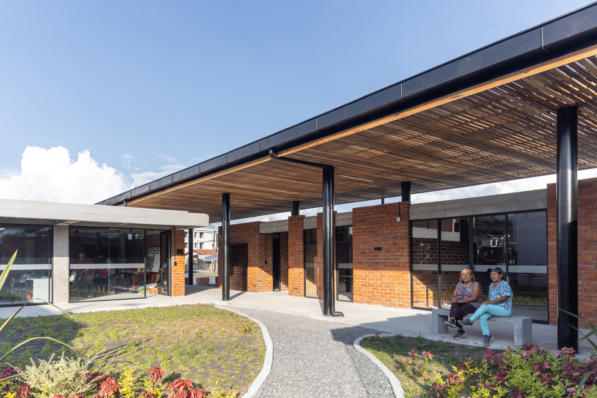 Covered porch with slatted wood ceiling supported by black columns overlooking a landscaped courtyard with seated visitors