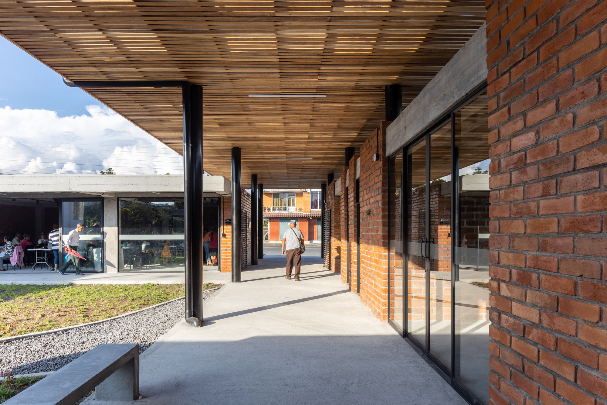 Linear covered walkway with timber ceiling, brick walls and glazed doors casting shadows under afternoon sun