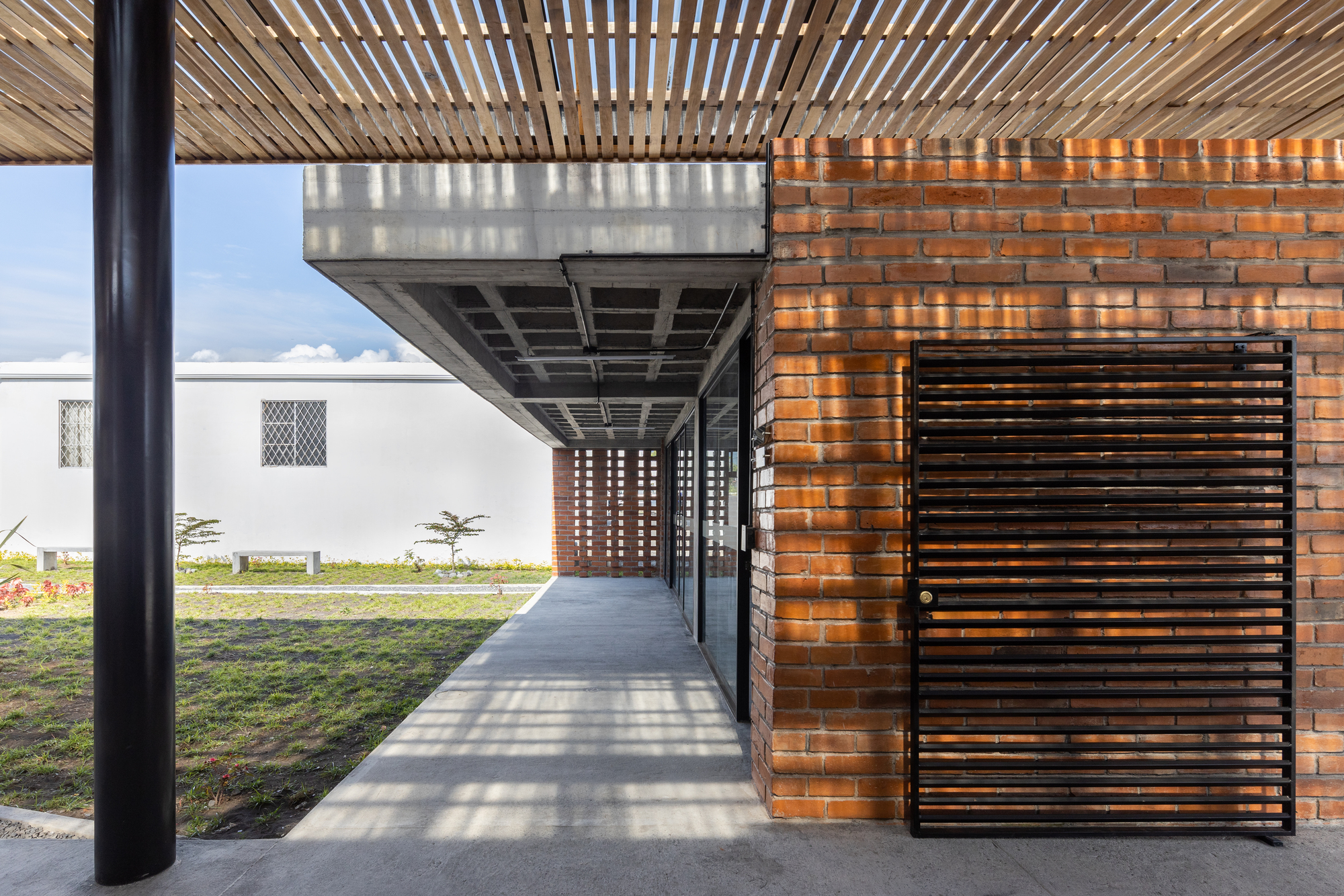 Exterior corridor with slatted timber ceiling, perforated brick screen and horizontal wood door casting dappled light