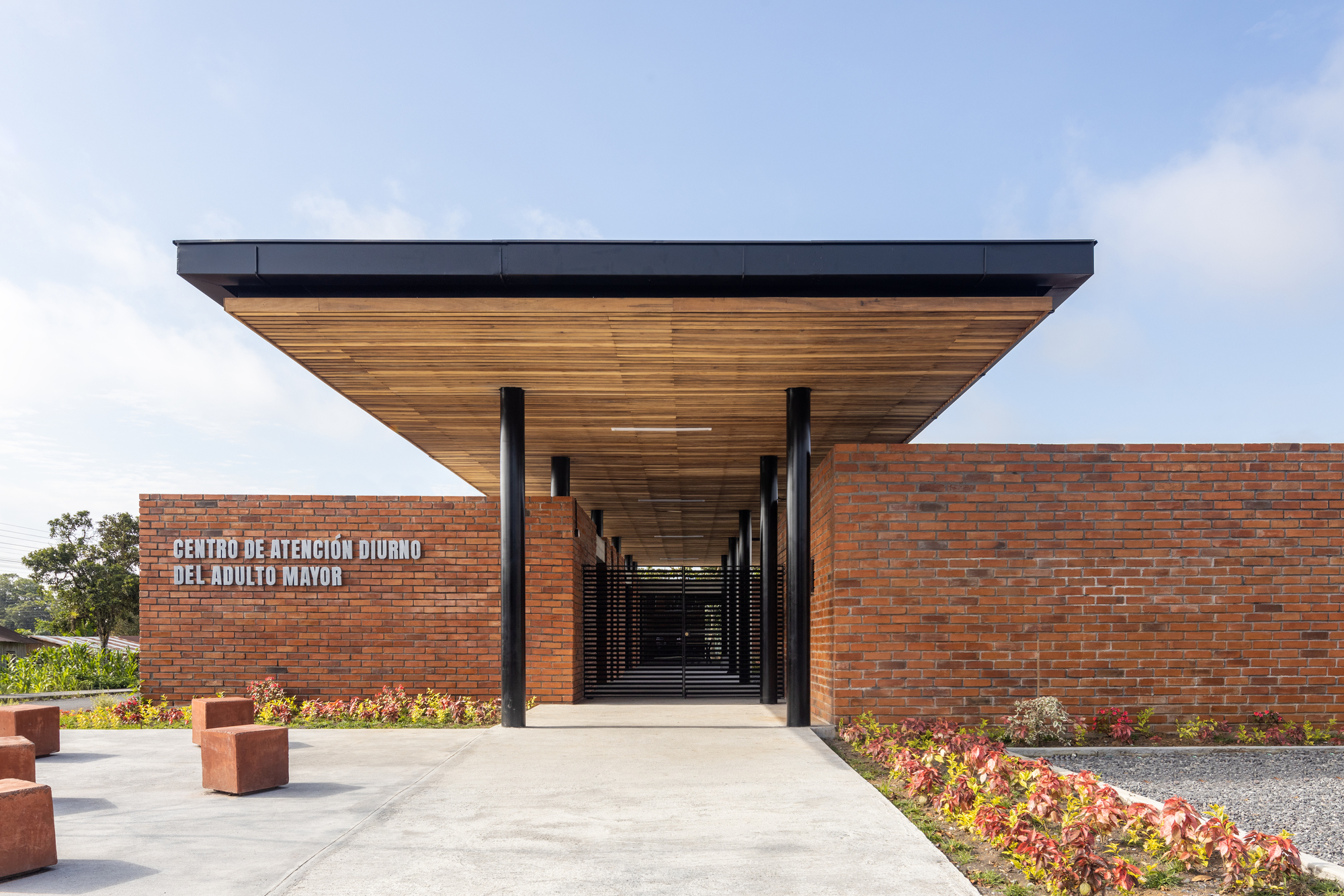 Entry facade with cantilevered timber soffit projecting over red brick wall and planted beds in daylight