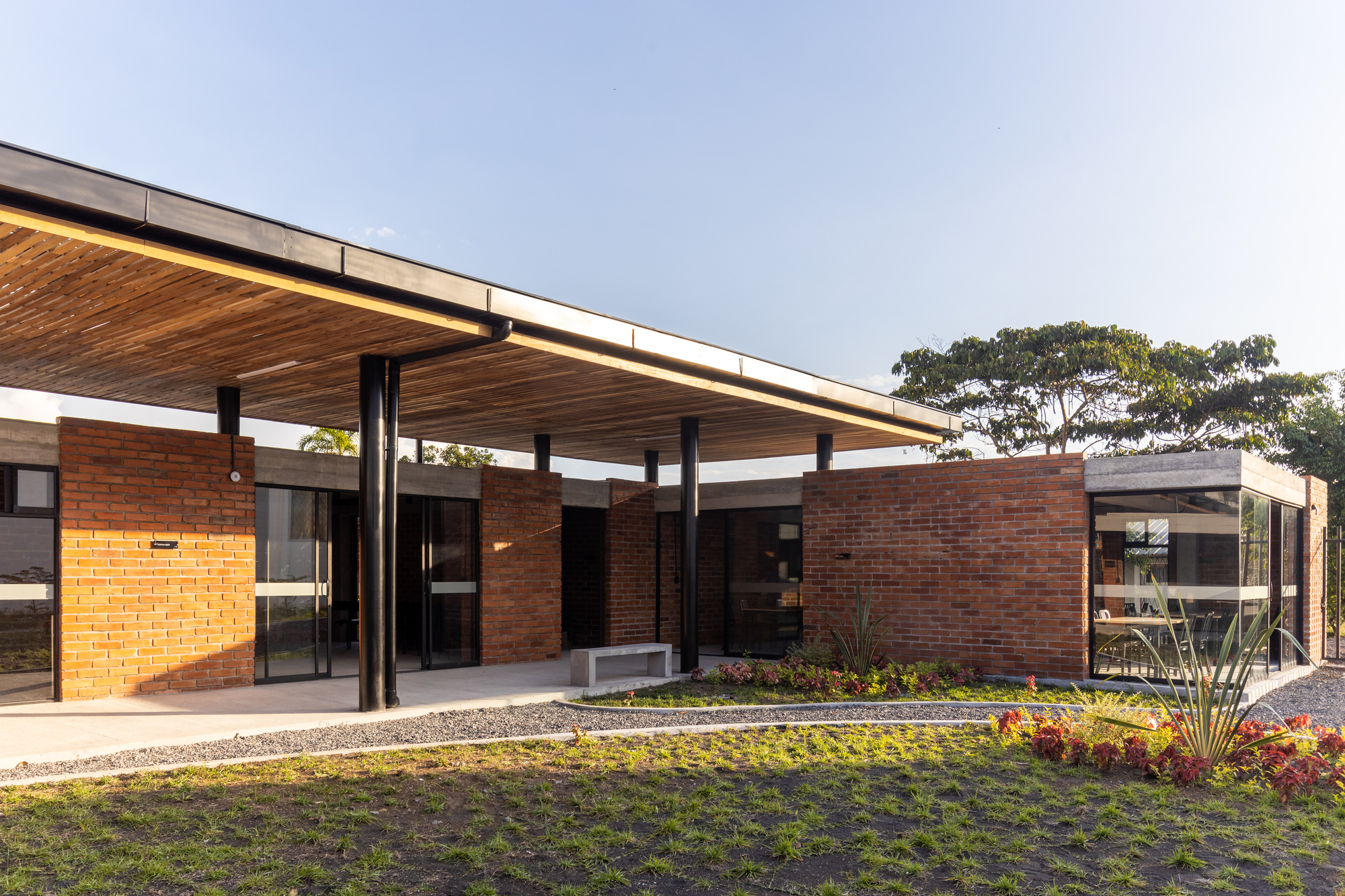Entry pavilion with extended timber slat roof, steel columns, brick walls, and planted beds at dusk