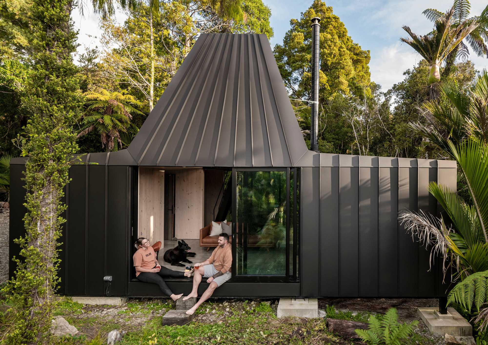 Black corrugated metal pavilion with conical roof and two people seated in open threshold beneath palms