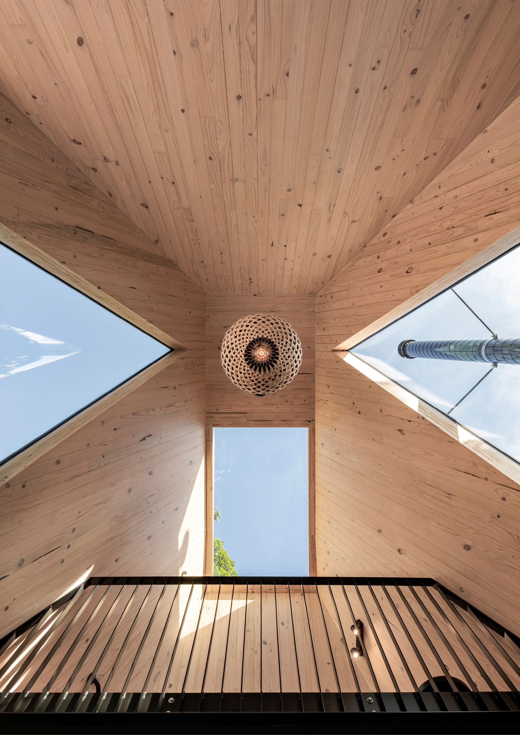 Upward view of timber-lined peaked ceiling with angled skylights and woven pendant centered above steel mezzanine railing