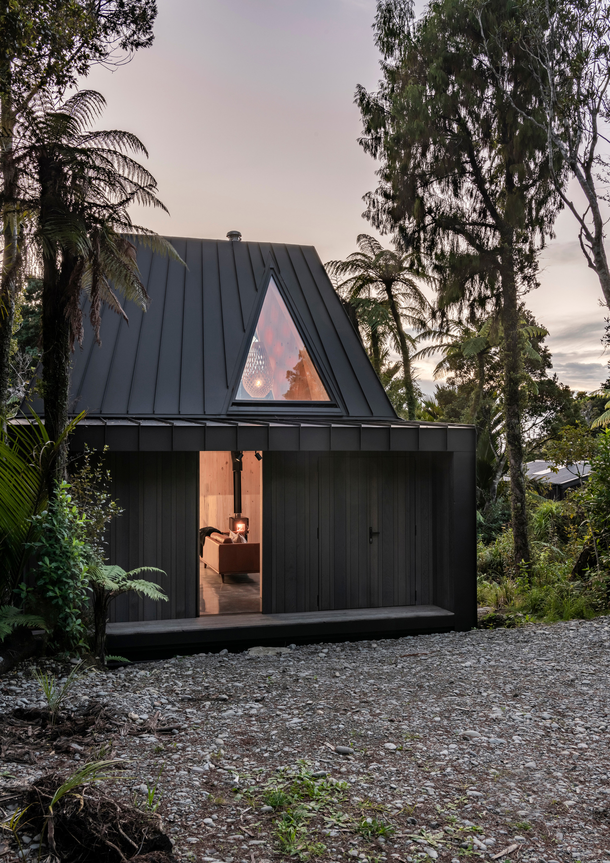Steep-pitched black metal roof with triangular window glowing at twilight among tropical trees