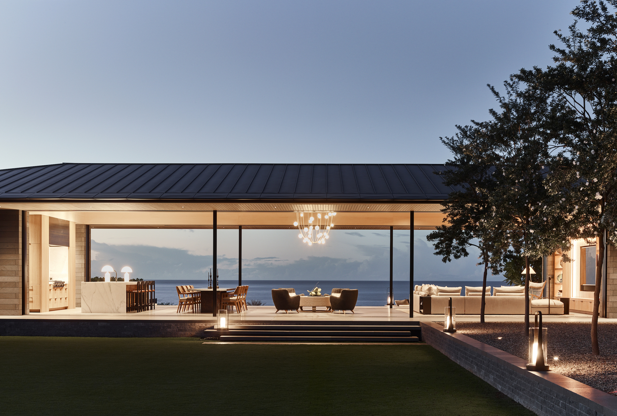 Glass-walled pavilion with standing seam metal roof opening to terrace and ocean view at twilight
