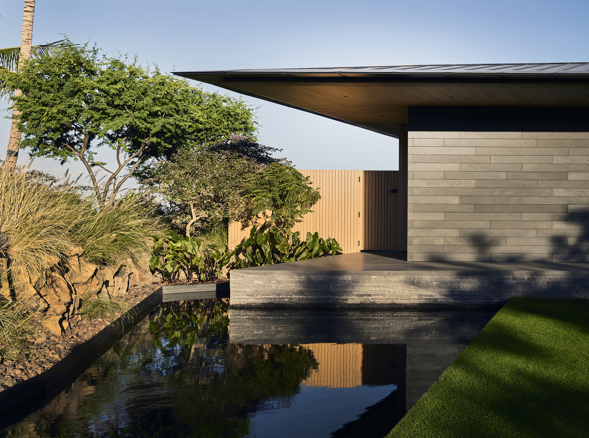 Entry courtyard with reflecting pool, horizontal stone wall, and cantilevered roof casting afternoon shadows