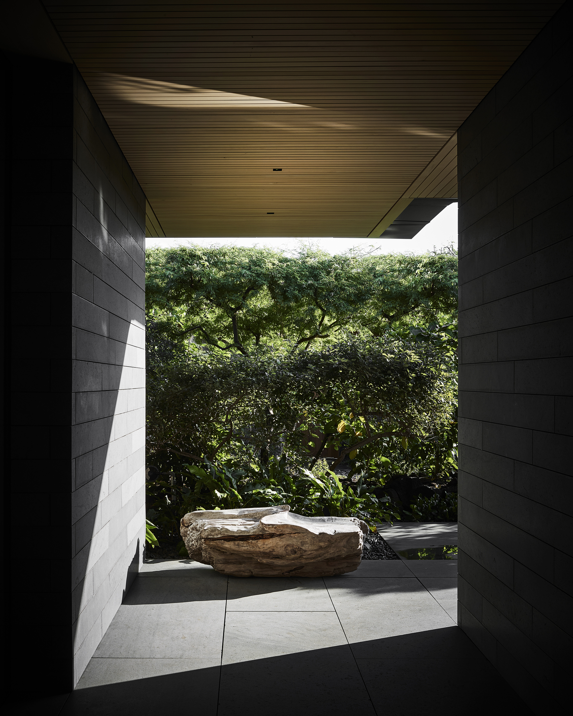Covered passage with ribbed wood ceiling framing a sculptural boulder and dense tree canopy beyond