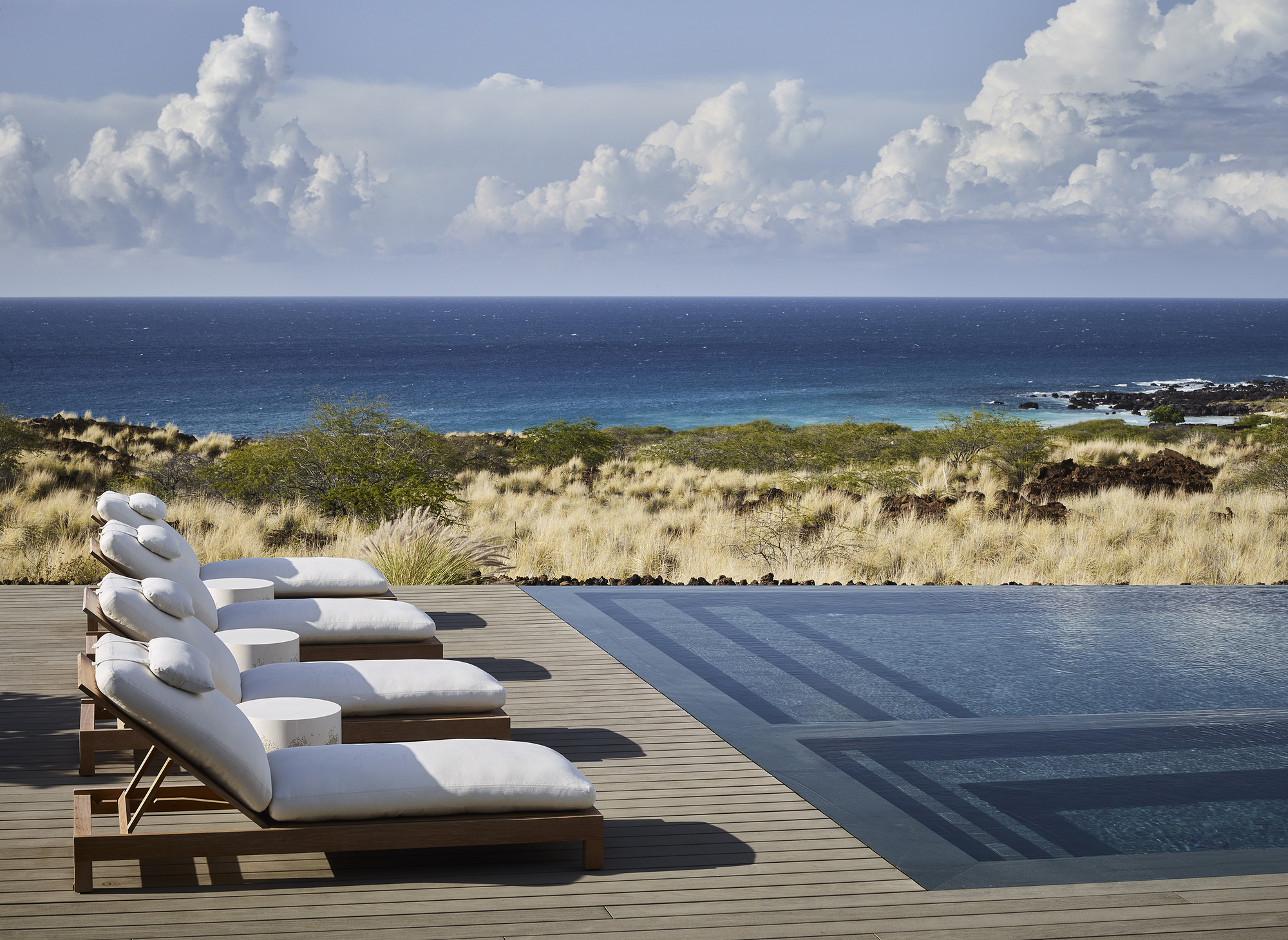 Timber deck with lounge chairs beside an infinity pool overlooking coastal grassland and ocean