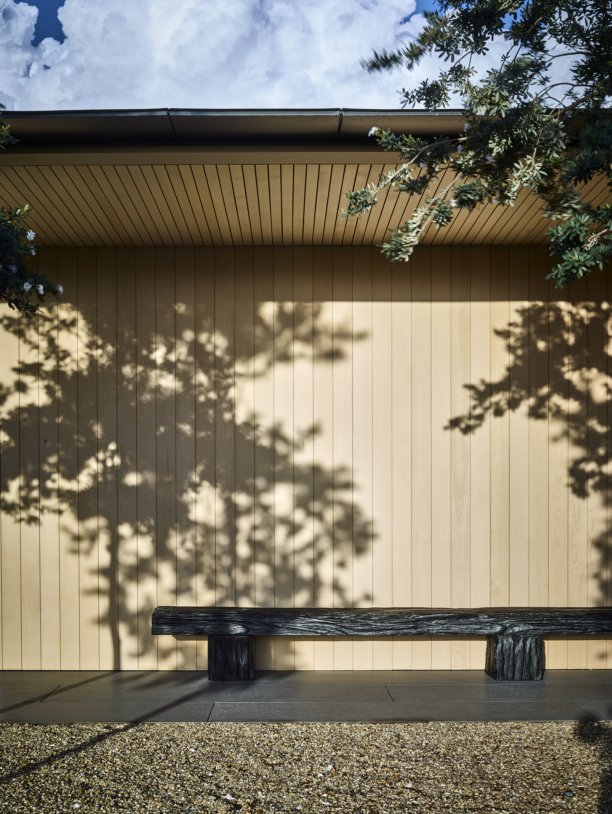 Vertical timber cladding with dappled tree shadow and charred wood bench on gravel