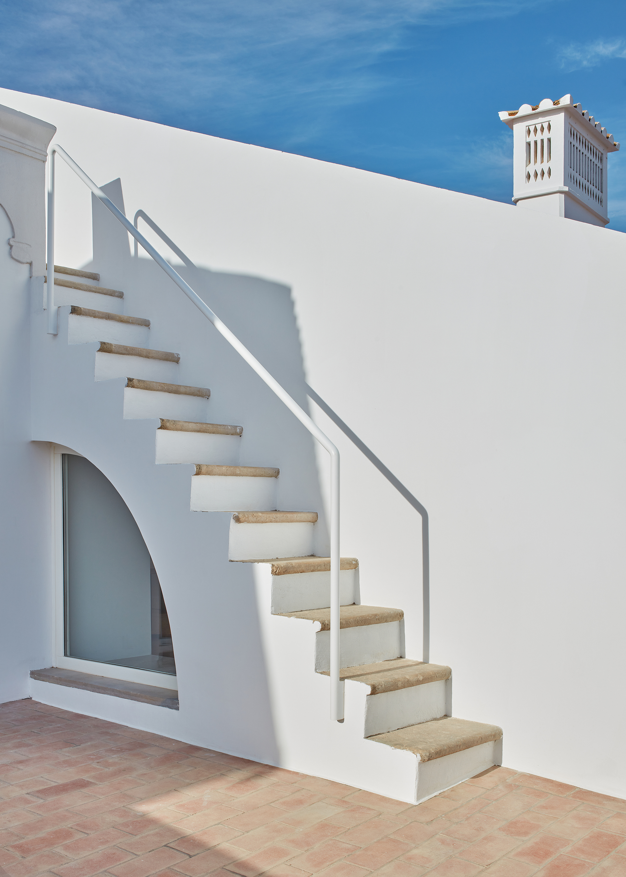 Exterior staircase with stone treads and white stucco wall casting sharp shadows on terracotta paving