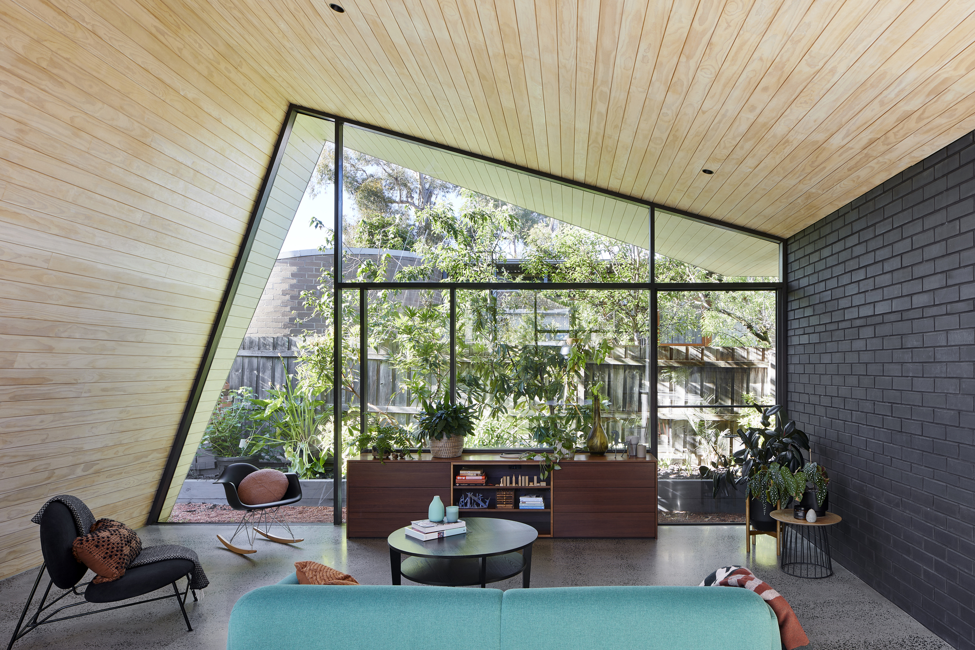 Living room with angled glazed wall framing garden views, timber plank ceiling and polished concrete floor