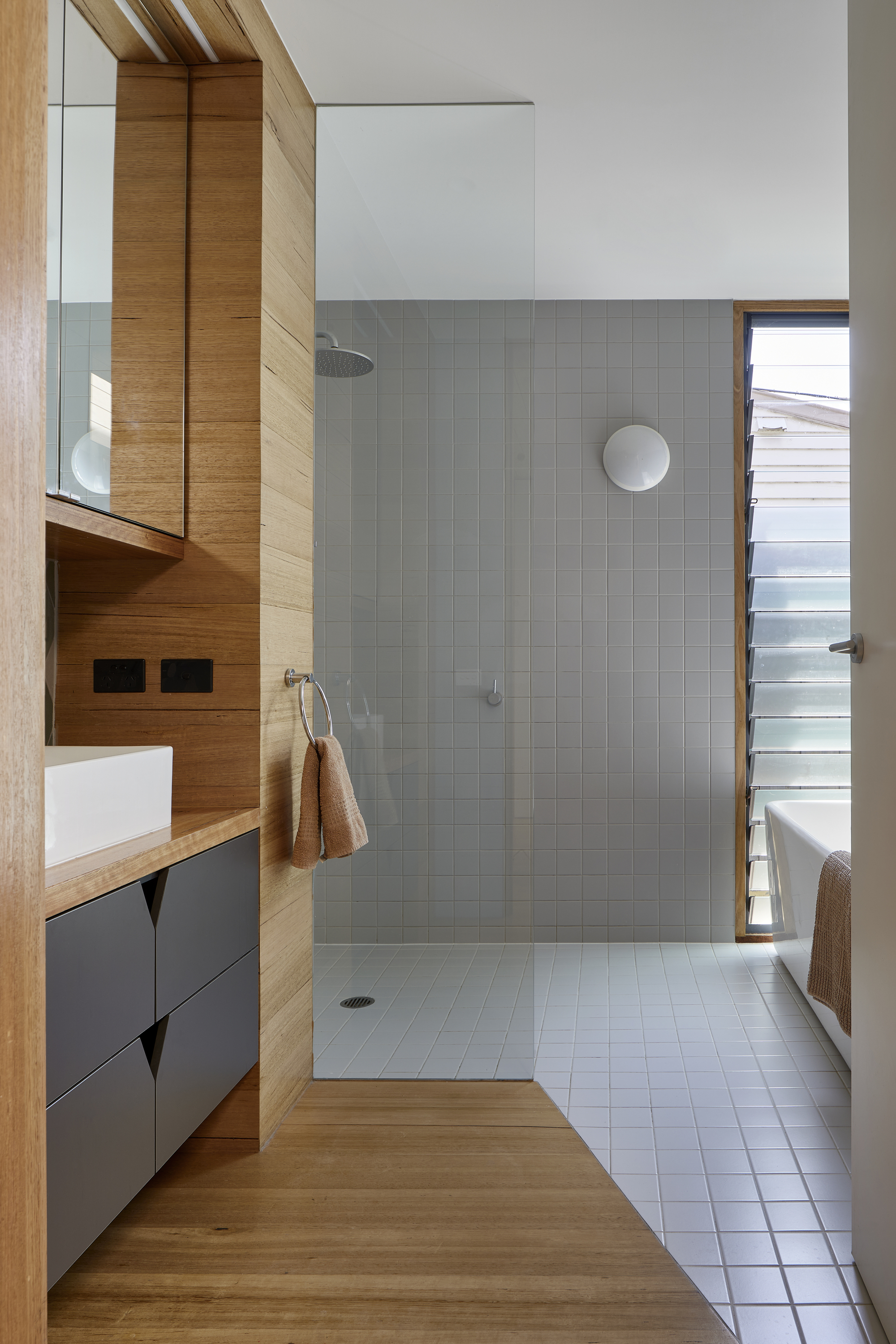 Bathroom with grey tile shower enclosure, timber vanity cabinetry and narrow glazed door to exterior