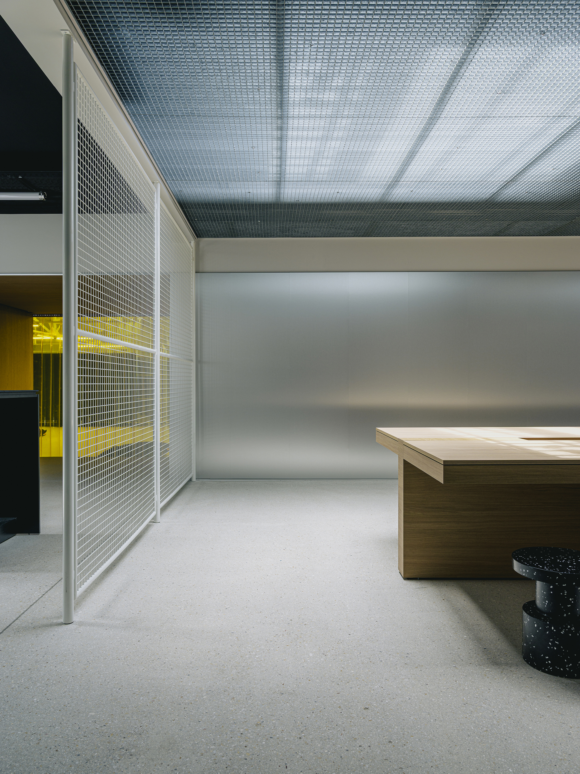 Interior space with metal mesh partition panels and yellow-lit room visible beyond timber desk