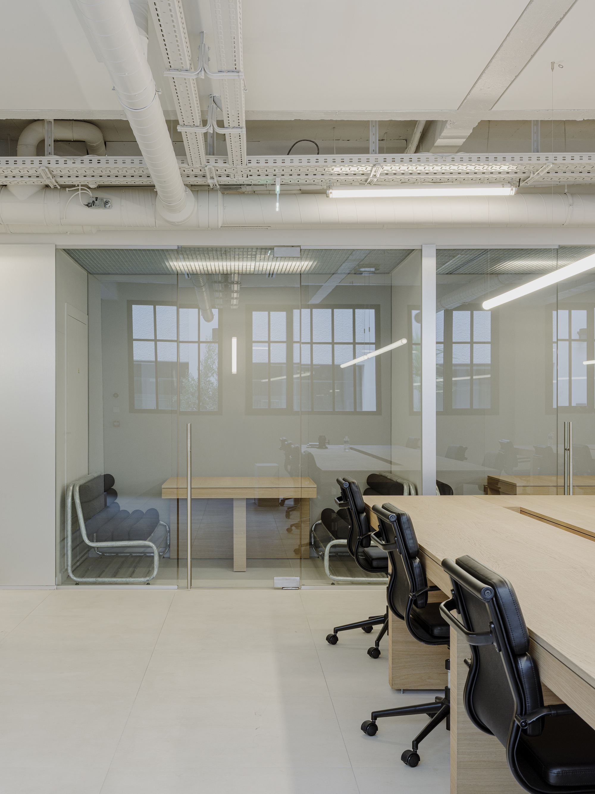 Open workspace with glass-enclosed meeting room, light wood desks, and exposed white structural beams overhead