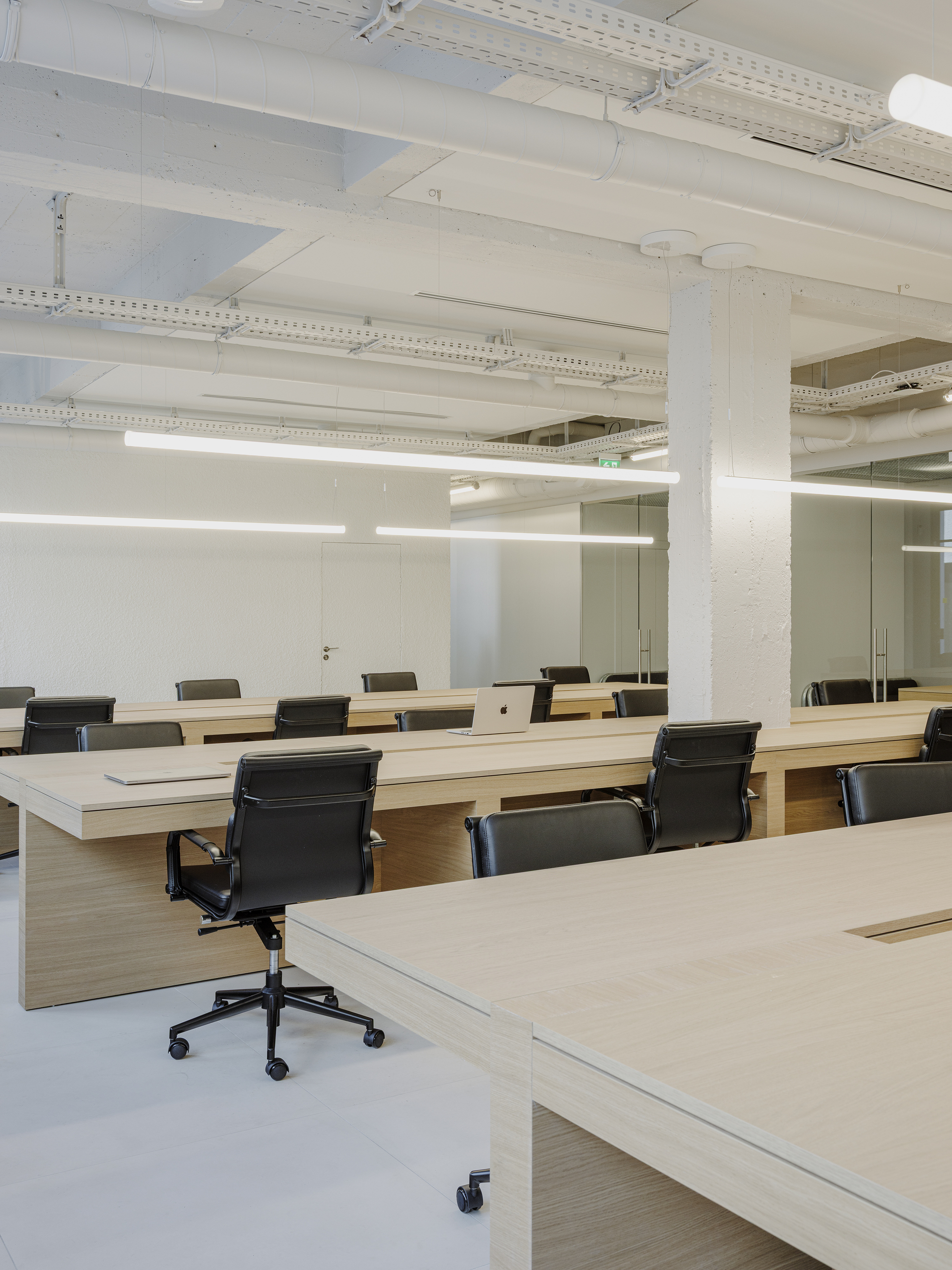 Rows of light wood desks with black office chairs under painted white structural beams and ductwork
