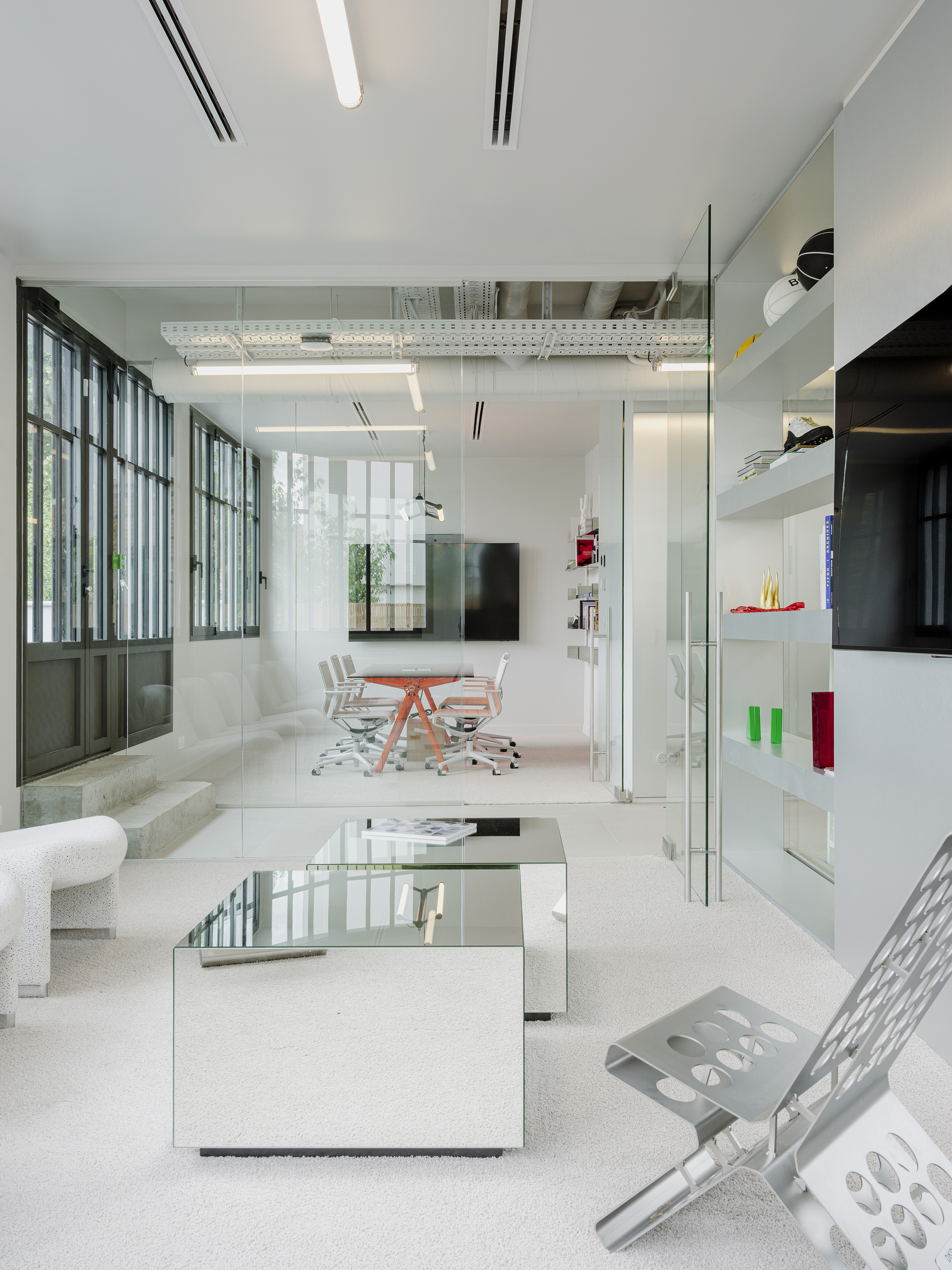 Double-height office space with glass partitions, mirrored coffee table, and steel-framed windows overlooking greenery