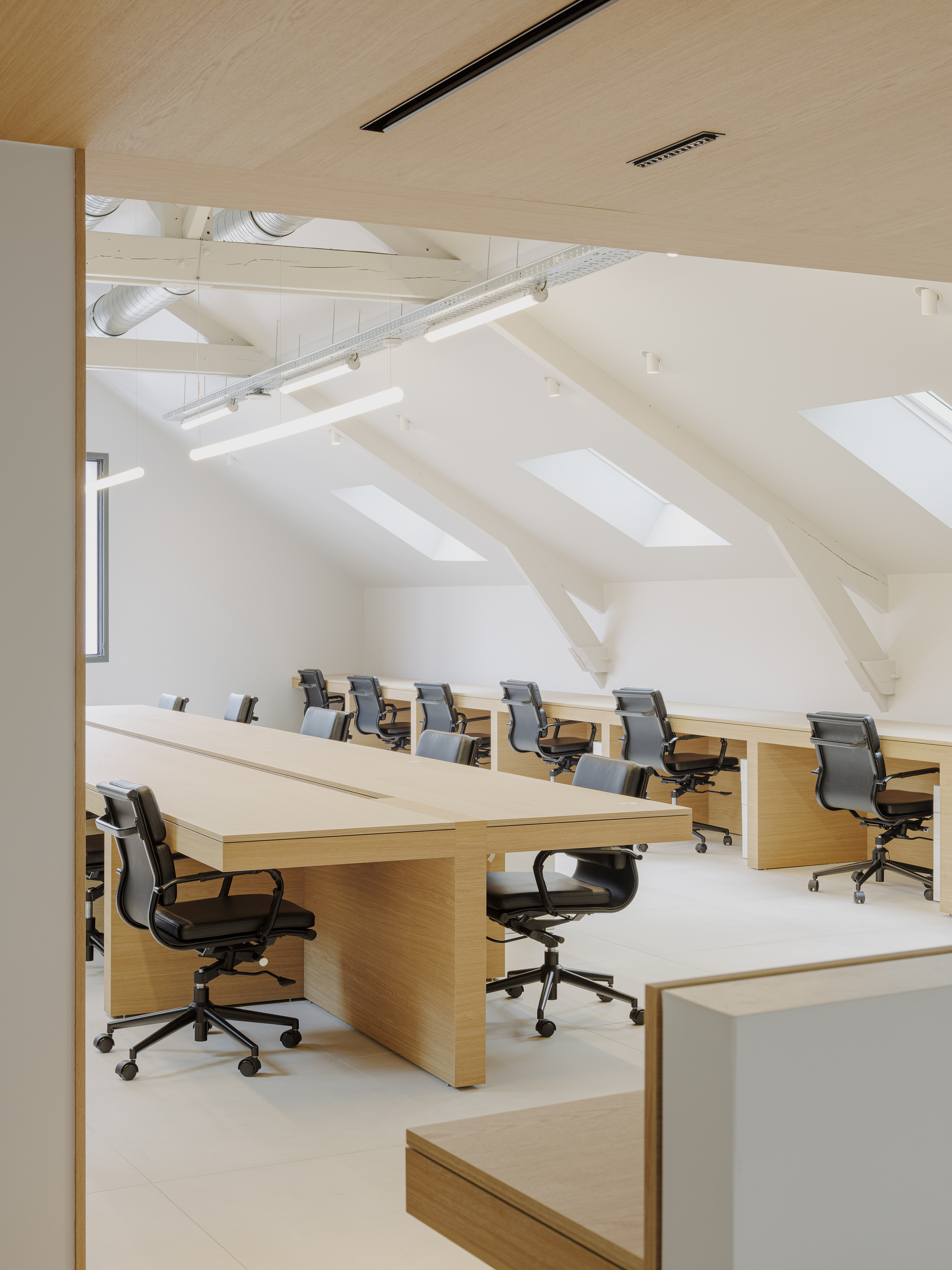 Open-plan office with rows of oak desks and office chairs under exposed white-painted roof trusses