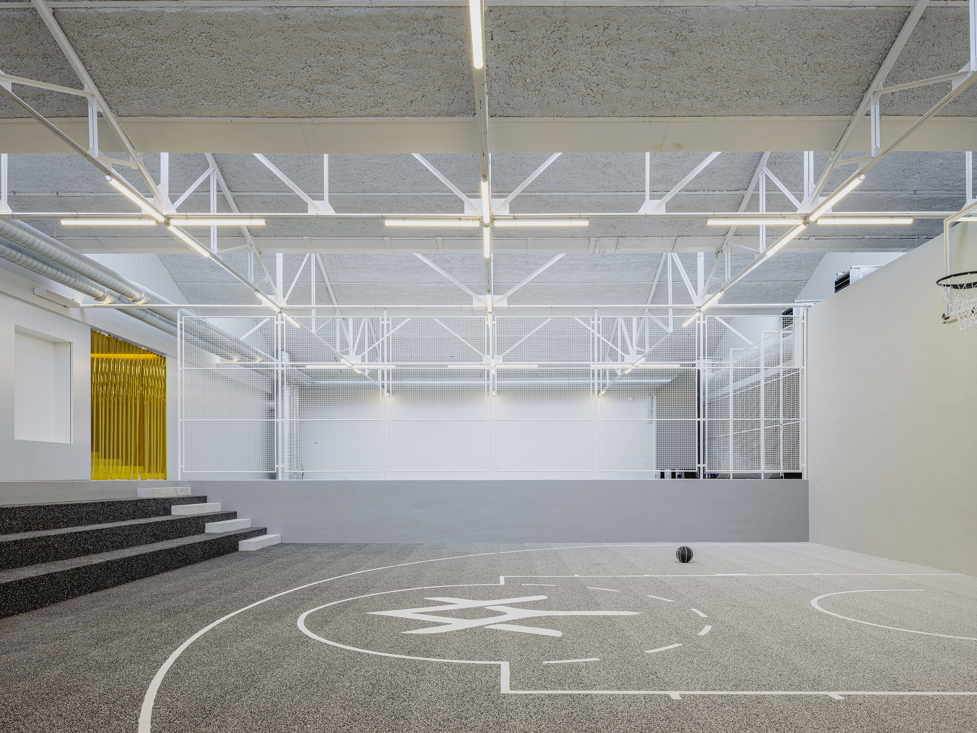 Gymnasium with white floor markings, exposed steel trusses, and tiered concrete seating along one wall
