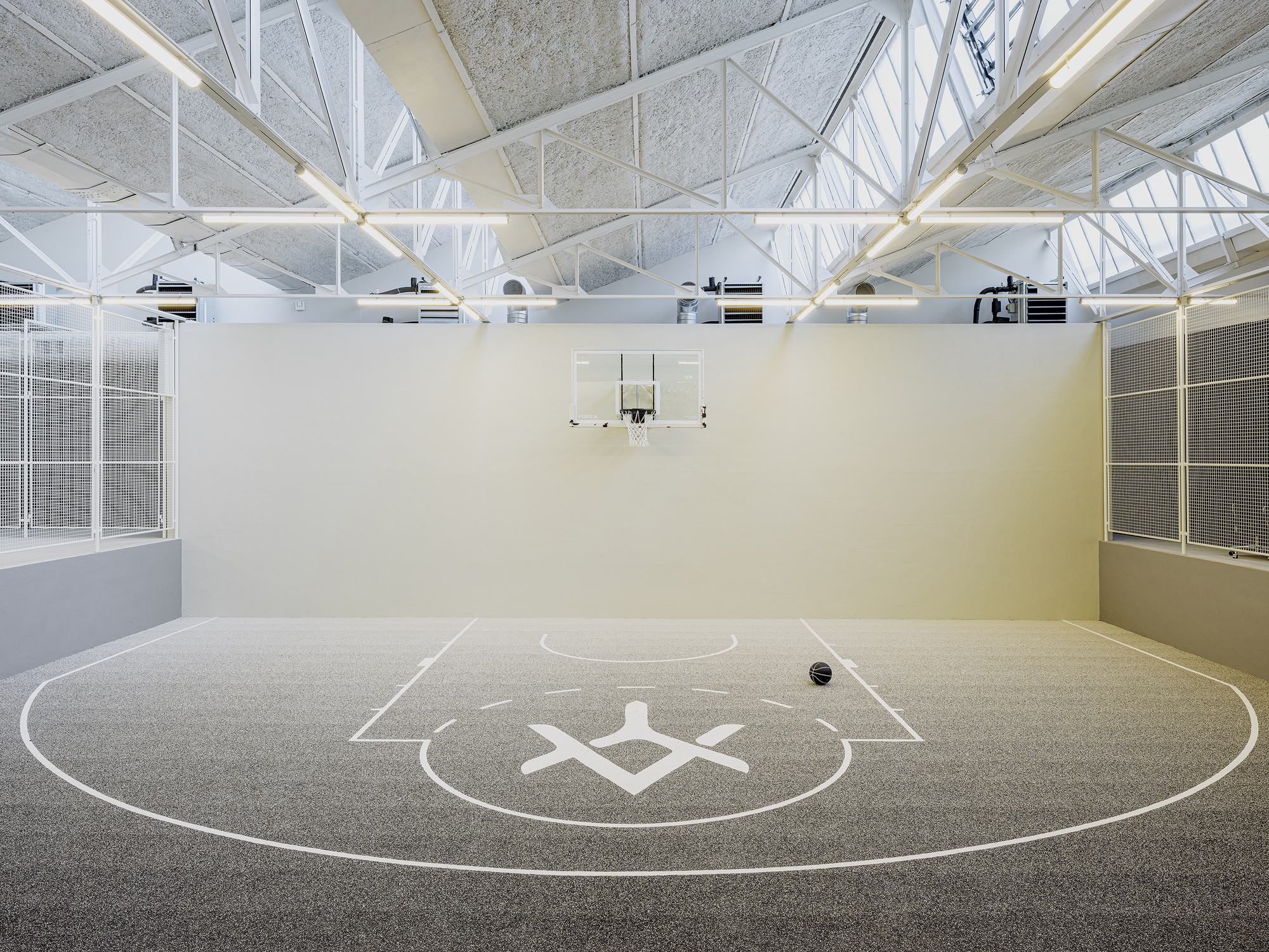 Indoor basketball court with white structural trusses overhead and tall glazed walls on both sides